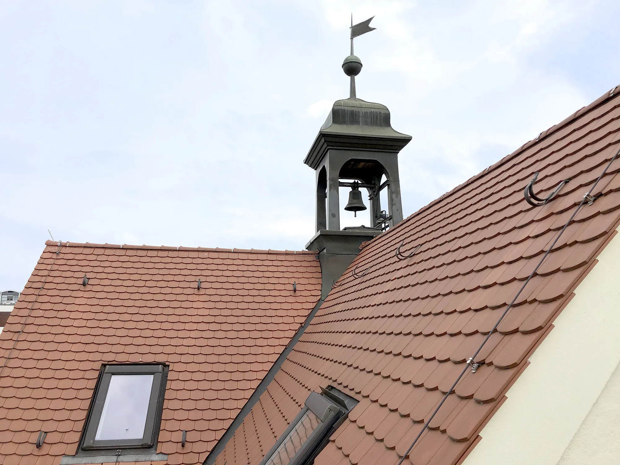 Dach eines Gebäudes mit roten Ziegeln und zwei Dachfenstern, im Hintergrund ein kleiner Turm mit einer Glocke und einer Wetterfahne unter leicht bewölktem Himmel.