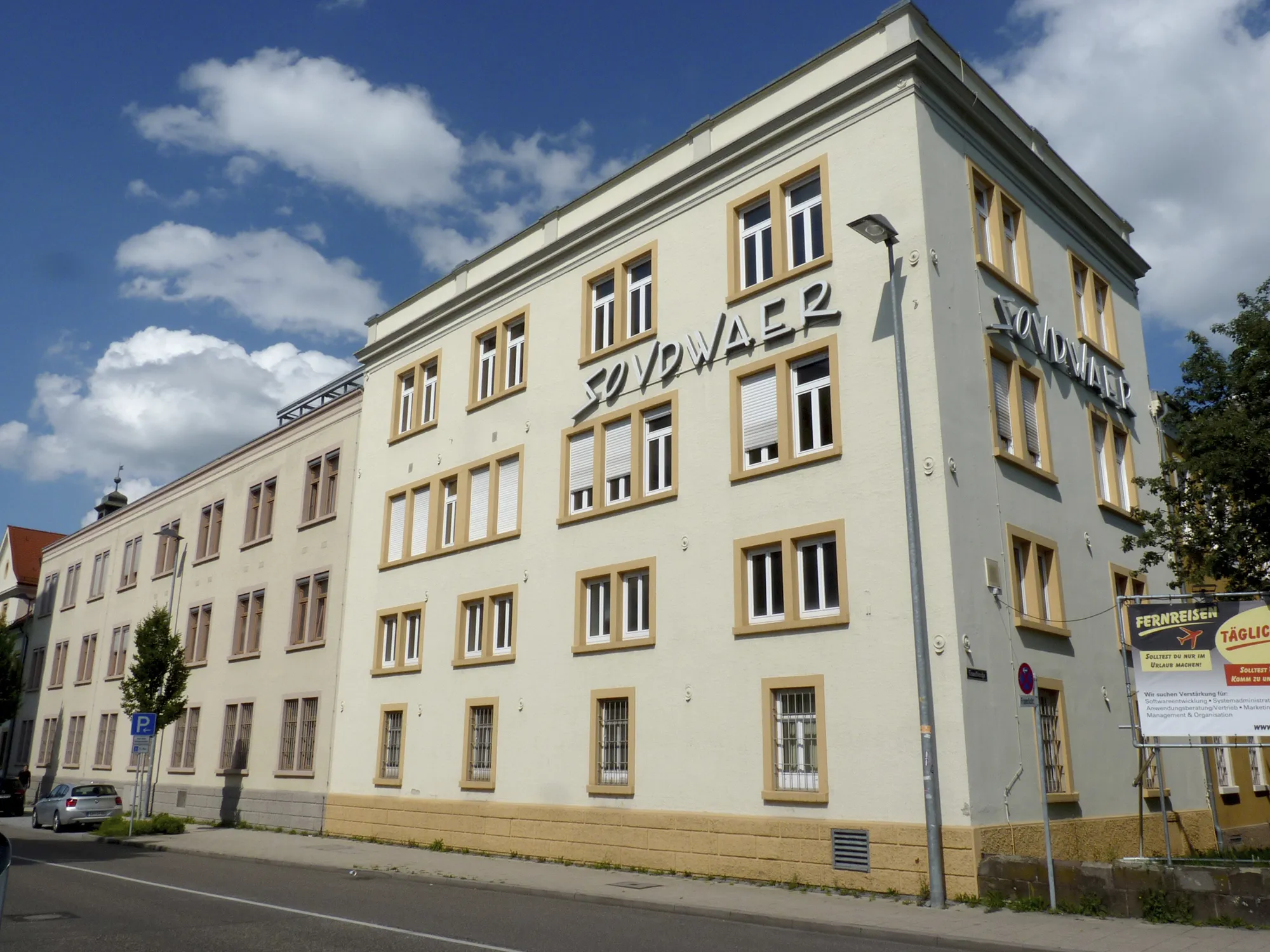 Ansicht eines hellen, mehrstöckigen Gebäudes mit gelb umrandeten Fenstern und dem Schriftzug „SOVDWAER“ an der Fassade, aufgenommen bei sonnigem Wetter mit blauem Himmel und einigen weißen Wolken.