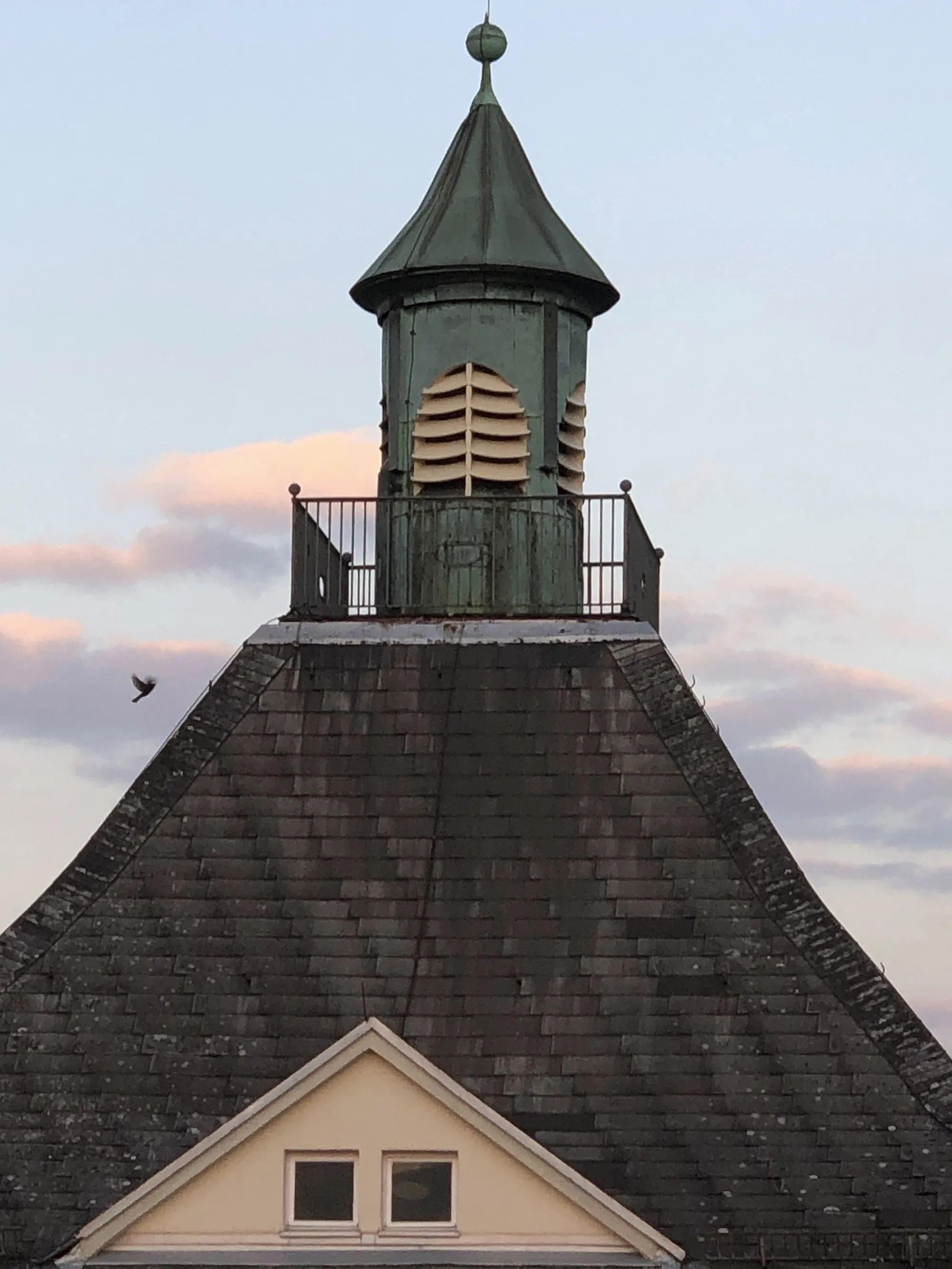 Altes, grün patiniertes Türmchen mit Kuppeldach und Lamellenfenstern, auf einem steilen Schieferdach montiert, umgeben von einer kleinen Plattform mit Geländer, vor einem Himmel mit rosa Wolken bei Sonnenuntergang.