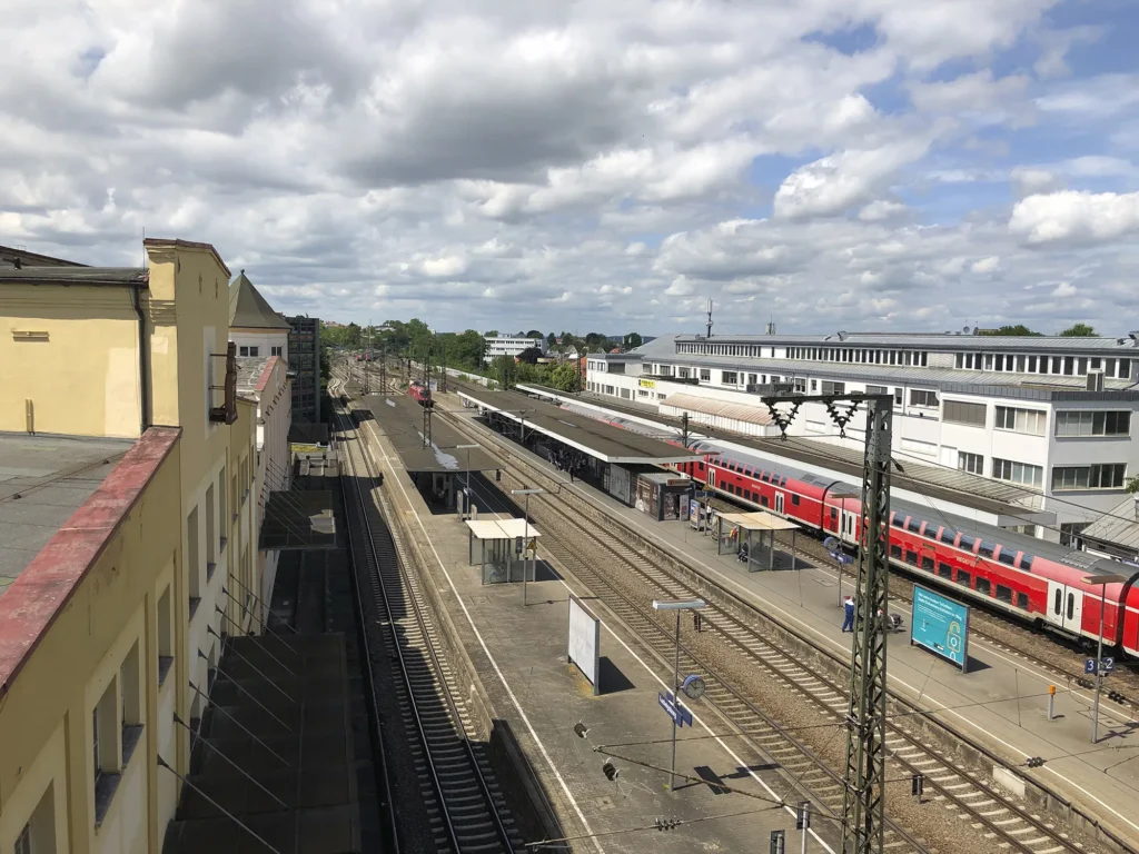 Blick von oben auf den Bahnhof Ludwigsburg mit mehreren Gleisen und überdachten Bahnsteigen, rechts hält ein roter Regionalzug, links die gelbe Fassade eines der Gebäude aus dem Caro Carré unter wechselnder Bewölkung.