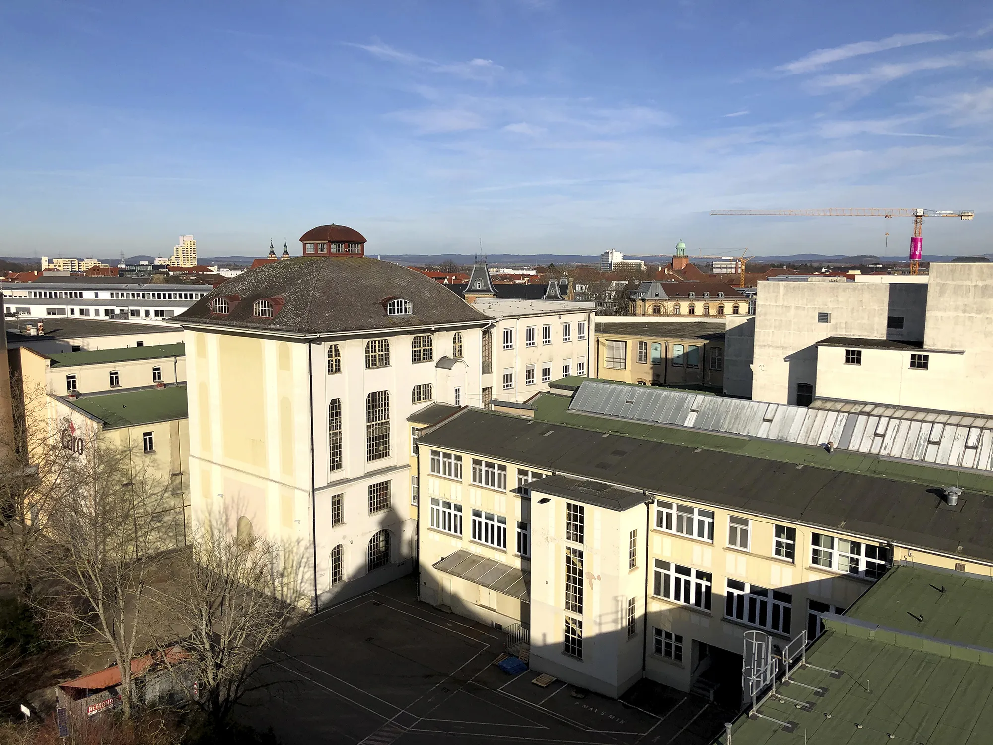 Blick über ein Industrie- und Bürogebäude-Ensemble mit einem großen, hellen Hauptgebäude mit gewölbtem Dach und vielen Fenstern, im Vordergrund weitere Gebäude mit grünen Dächern, im Hintergrund die Skyline von Ludwigsburg mit Kran, Türmen und klarem blauen Himmel.