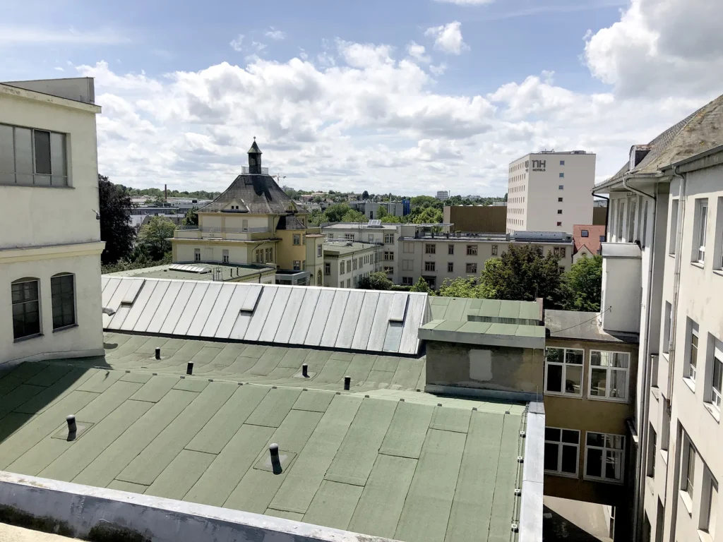 Blick über mehrere Gebäudedächer, im Vordergrund grüne und graue Flachdächer, im Mittelgrund ein großes gelbes Gebäude mit markantem Turm und Schieferdach, im Hintergrund Bäume, weitere Häuser und ein Hotelhochhaus unter teils bewölktem Himmel.