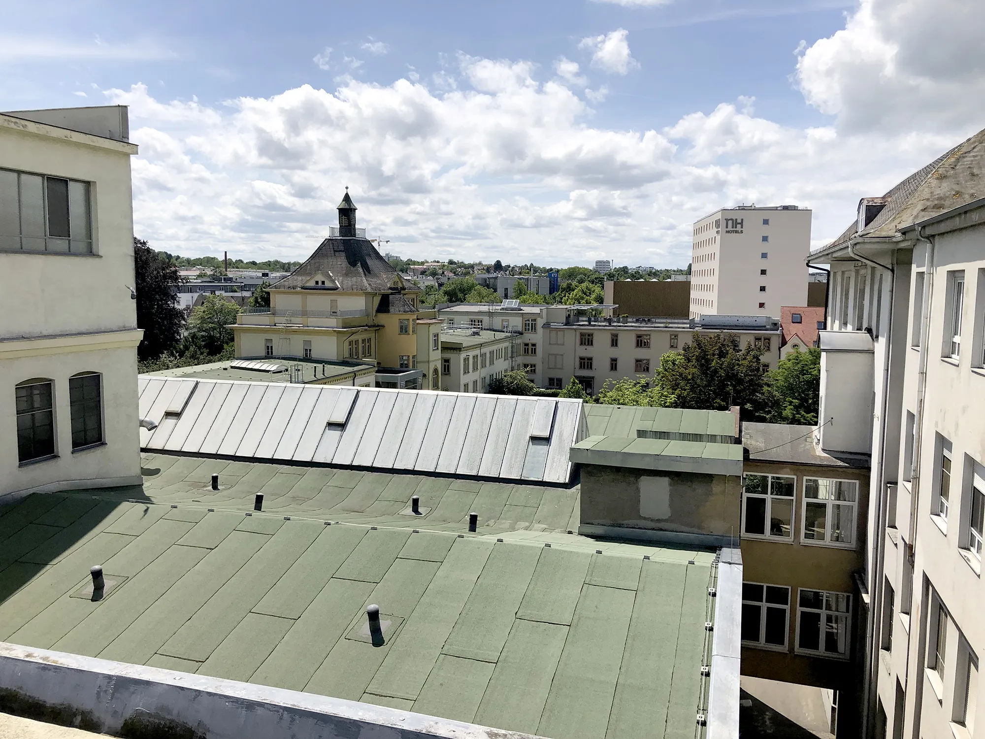 Blick über mehrere Gebäudedächer, im Vordergrund grüne und graue Flachdächer, im Mittelgrund ein großes gelbes Gebäude mit markantem Turm und Schieferdach, im Hintergrund Bäume, weitere Häuser und ein Hotelhochhaus unter teils bewölktem Himmel.
