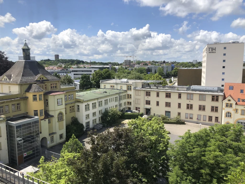 Luftaufnahme des Caro Carrés inklusive Innenhof. Im Vordergrund große grüne Bäume, im Hintergrund ein Hotelgebäude sowie viele andere Gebäude.
