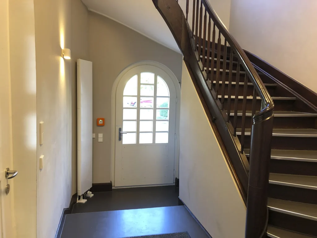 Eingangsbereich mit weißer, halb verglaster Tür in Rundbogenform, daneben eine Wandlampe und ein roter Notfallknopf, rechts eine hölzerne Treppe mit dunklem Handlauf, die nach oben führt.