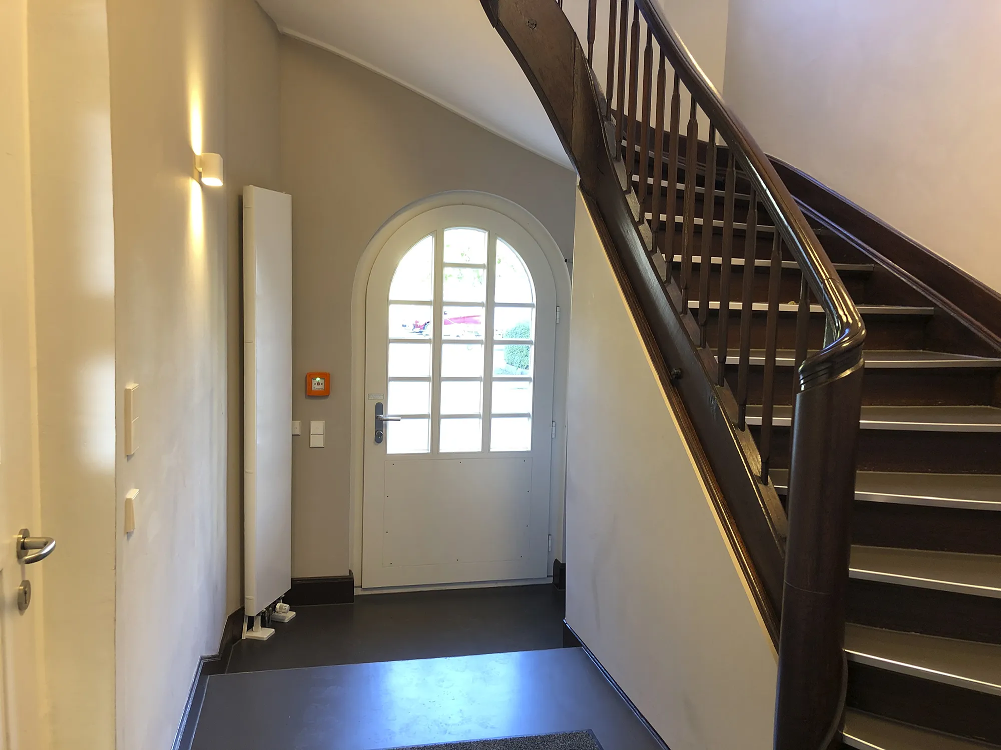 Eingangsbereich mit weißer, halb verglaster Tür in Rundbogenform, daneben eine Wandlampe und ein roter Notfallknopf, rechts eine hölzerne Treppe mit dunklem Handlauf, die nach oben führt.