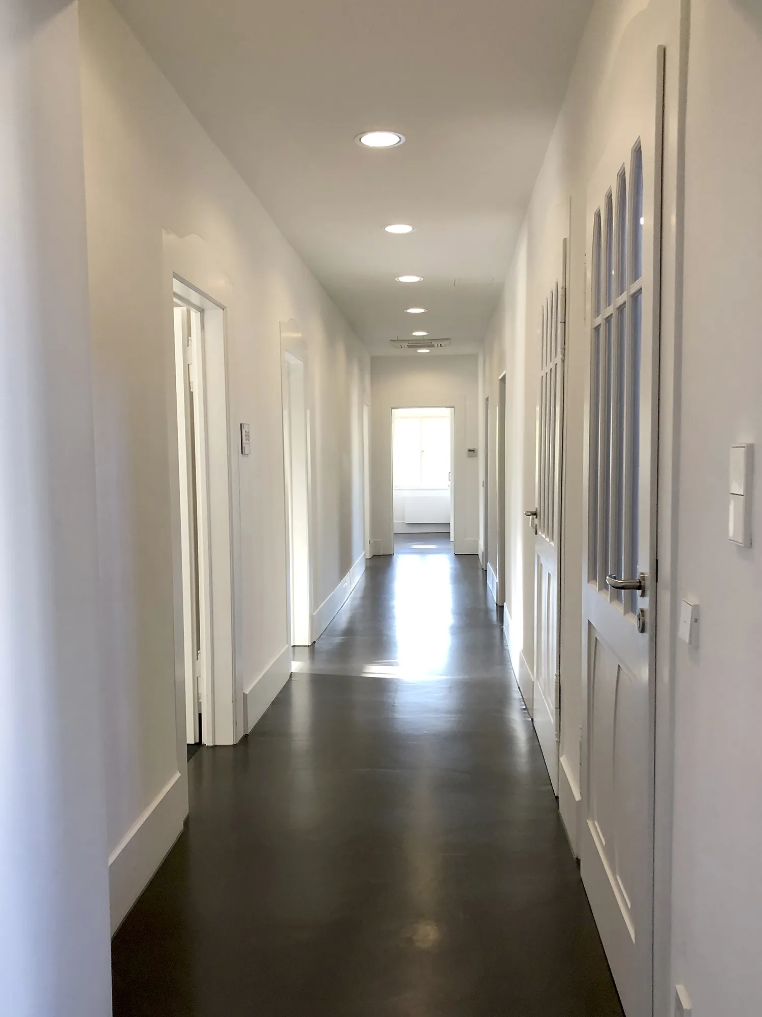 Langer, schmaler Flur mit weißen Wänden und Türen, dunklem glänzenden Boden und eingelassenen Deckenleuchten, am Ende ein Fenster, das Tageslicht hereinlässt.