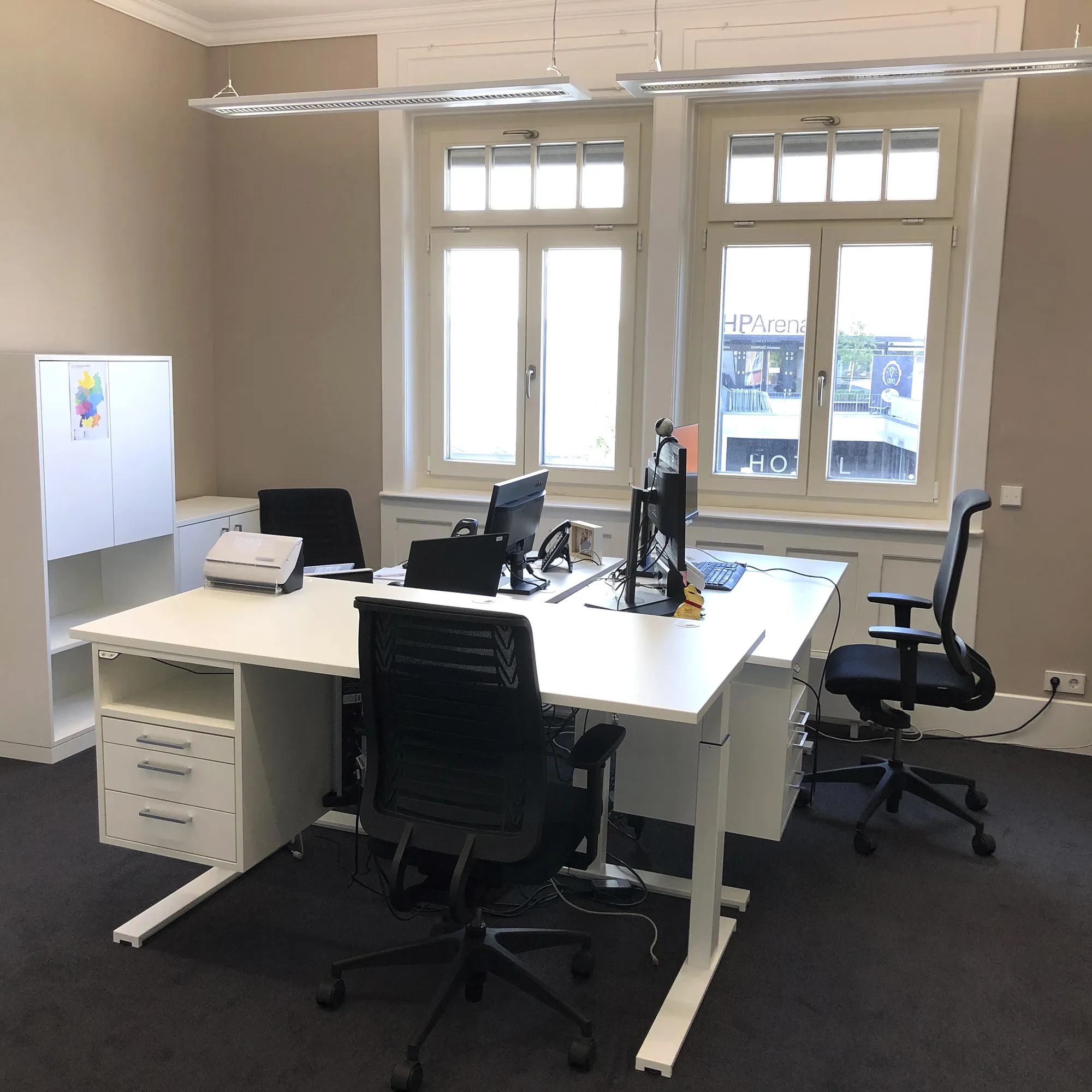 Modernes Büro mit zwei weißen Schreibtischen, schwarzen Bürostühlen und Computerarbeitsplätzen, davor zwei große Fenster mit Blick nach draußen, links ein weißer Aktenschrank.