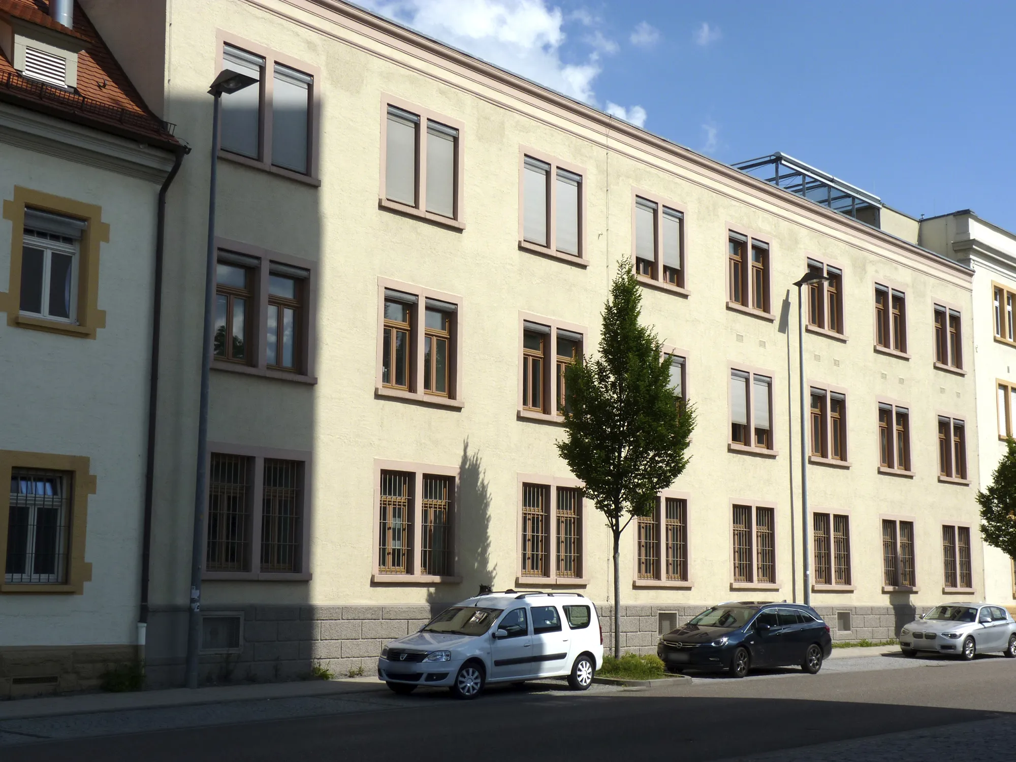 Dreigeschossige Straßenfassade eines hell verputzten Gebäudes, welches Teil vom Caro Carré ist, mit hochrechteckigen Fenstern und gegliedertem Sockel, davor Straßenbäume und geparkte Autos bei klarem Himmel.