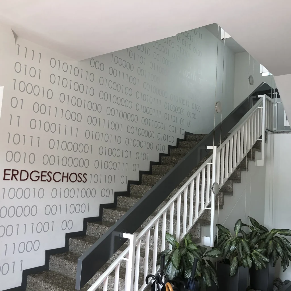 Innenansicht eines Treppenhauses mit grauen Stufen und weißem Geländer, flankiert von einer Wand mit großem Binärcode-Muster in Grau. An der Wand steht in roten Buchstaben „ERDGESCHOSS“. Unten am Treppenaufgang befinden sich mehrere große grüne Topfpflanzen.