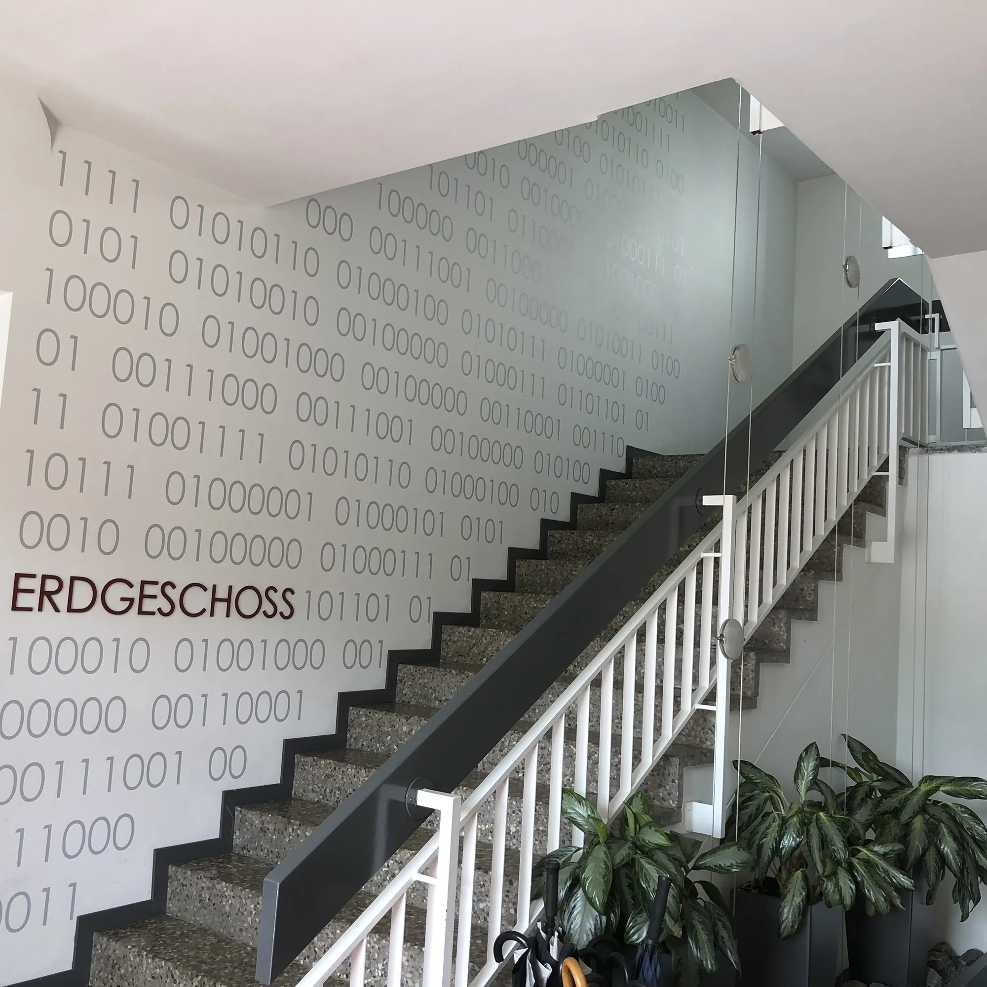 Innenansicht eines Treppenhauses mit grauen Stufen und weißem Geländer, flankiert von einer Wand mit großem Binärcode-Muster in Grau. An der Wand steht in roten Buchstaben „ERDGESCHOSS“. Unten am Treppenaufgang befinden sich mehrere große grüne Topfpflanzen.