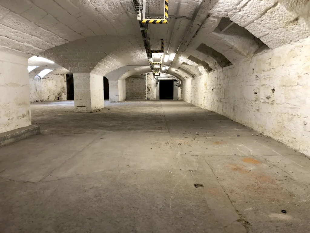 Langer, gewölbter Kellerraum mit weiß gestrichenen Steinwänden und mehreren Säulen, Betonboden und Deckenbeleuchtung.