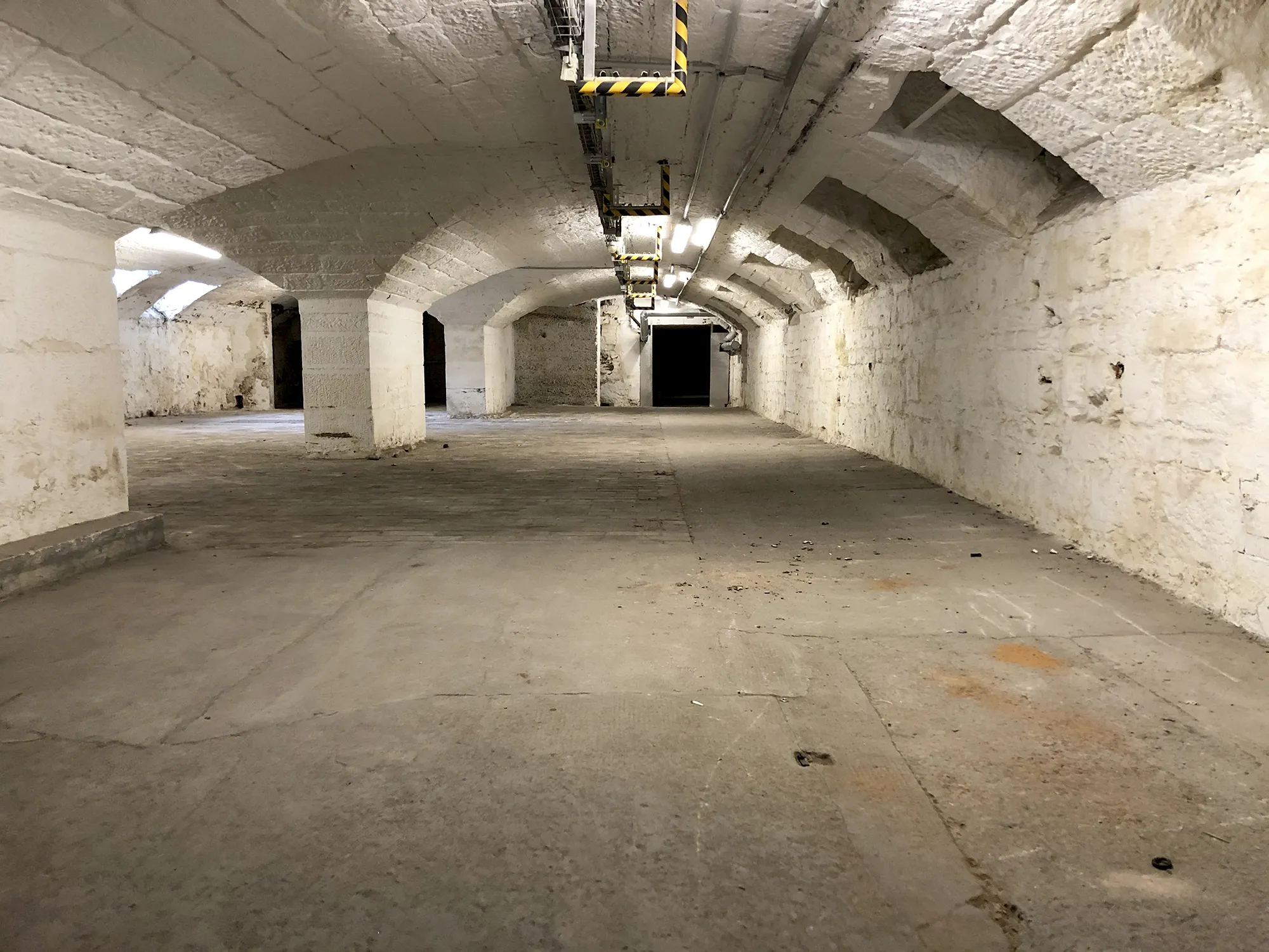 Langer, gewölbter Kellerraum mit weiß gestrichenen Steinwänden und mehreren Säulen, Betonboden und Deckenbeleuchtung.