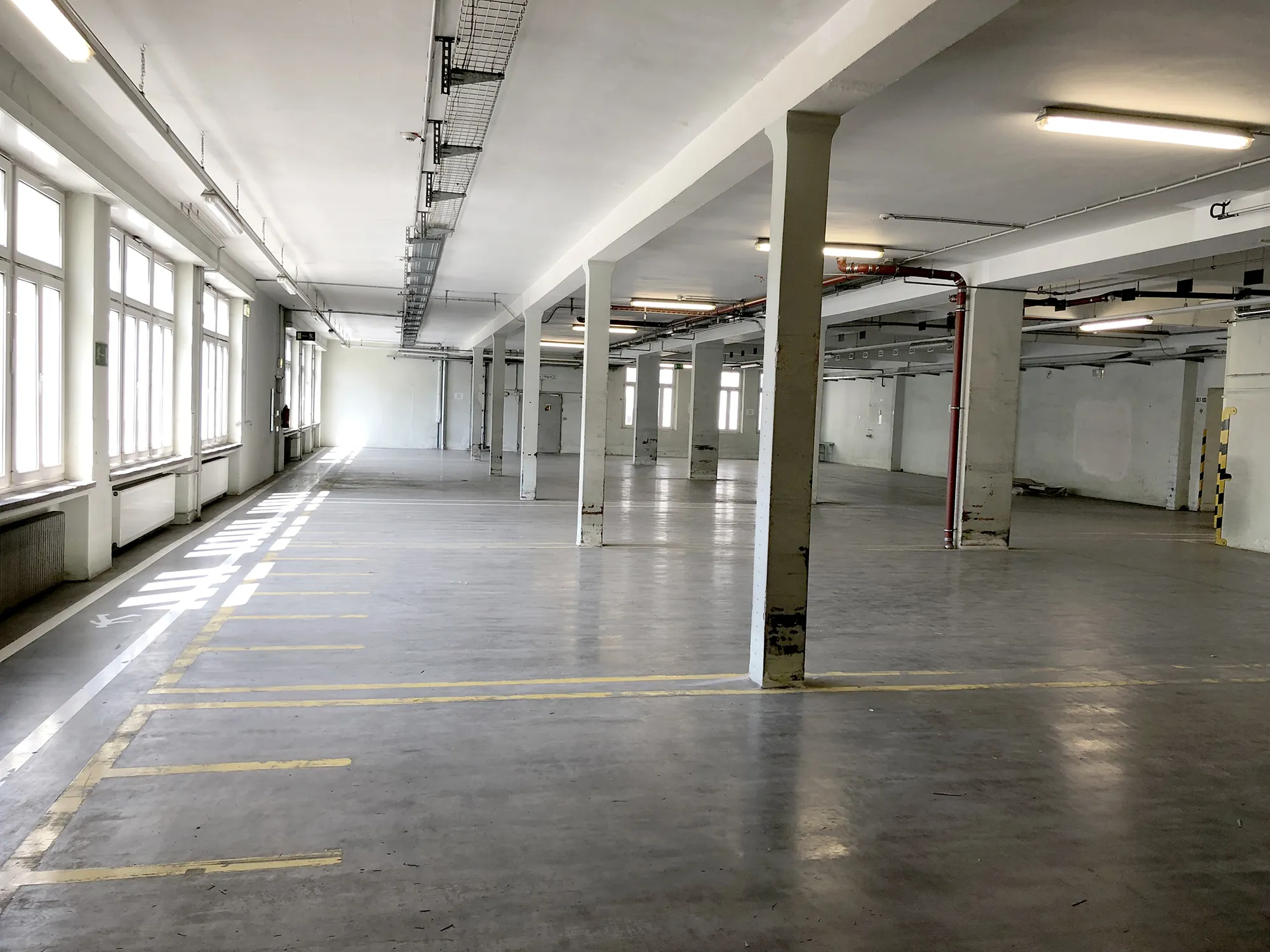Leere Industriehalle in einem Gebäude des caro Carrés mit Reihen von Betonsäulen, großen Fensterfronten links, Bodenmarkierungen und Deckenleuchten.