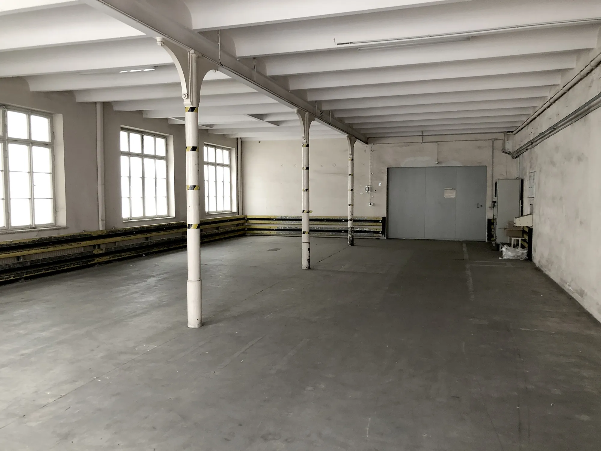 Leerer Industrie- oder Lagerraum mit großen Fenstern an der linken Seite, weiß gestrichenen Stützsäulen mit gelb-schwarzen Markierungen und einer großen grauen Doppeltür an der Stirnseite.