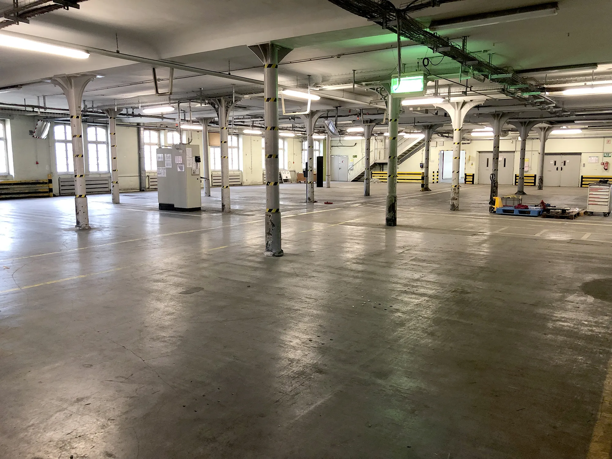 Große, leere Industriehalle mit zahlreichen weißen Stützsäulen, gelb-schwarzen Sicherheitsmarkierungen, Fenstern an den Seiten und einer Treppe im Hintergrund.
