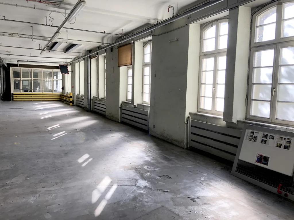 Leerer, lichtdurchfluteter Industriebereich mit großen Bogenfenstern, grauen Heizkörpern und einer verglasten Kabine im Hintergrund,.