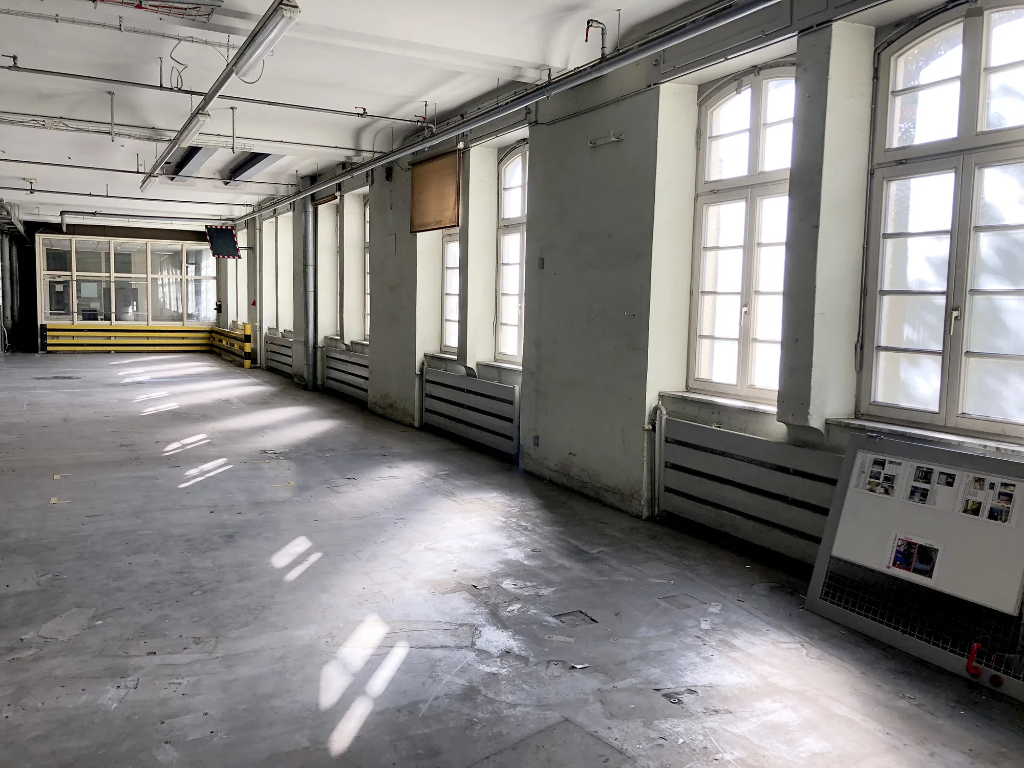 Leerer, lichtdurchfluteter Industriebereich mit großen Bogenfenstern, grauen Heizkörpern und einer verglasten Kabine im Hintergrund,.
