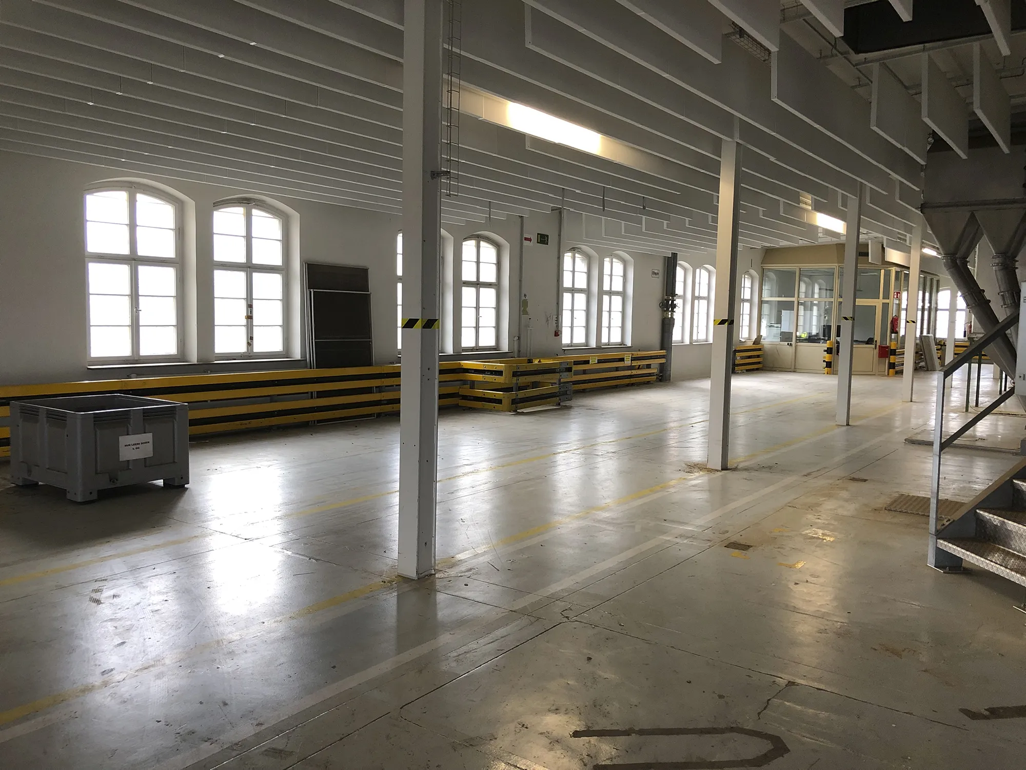 Großer, leerer Industrie- oder Lagerraum mit weißen Wänden, hohen Bogenfenstern, gelb-schwarzen Schutzbarrieren und einer verglasten Bürokabine im Hintergrund.