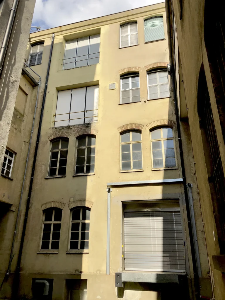Innenhofansicht eines mehrstöckigen Gebäudes mit gelber Fassade, mehreren Fenstern in unterschiedlichen Größen und Formen, sowie einem großen Rolltor im Erdgeschoss.
