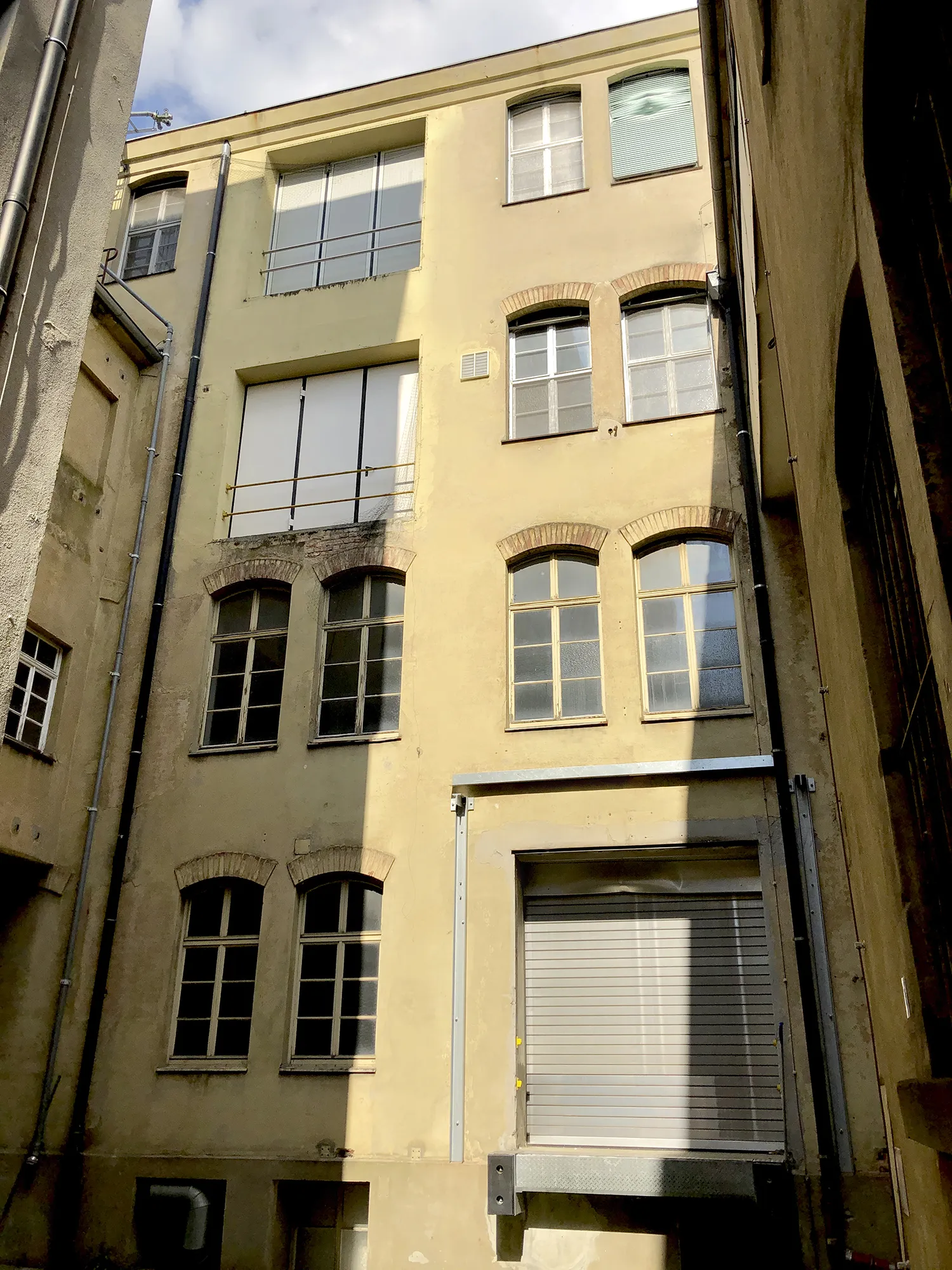 Innenhofansicht eines mehrstöckigen Gebäudes mit gelber Fassade, mehreren Fenstern in unterschiedlichen Größen und Formen, sowie einem großen Rolltor im Erdgeschoss.