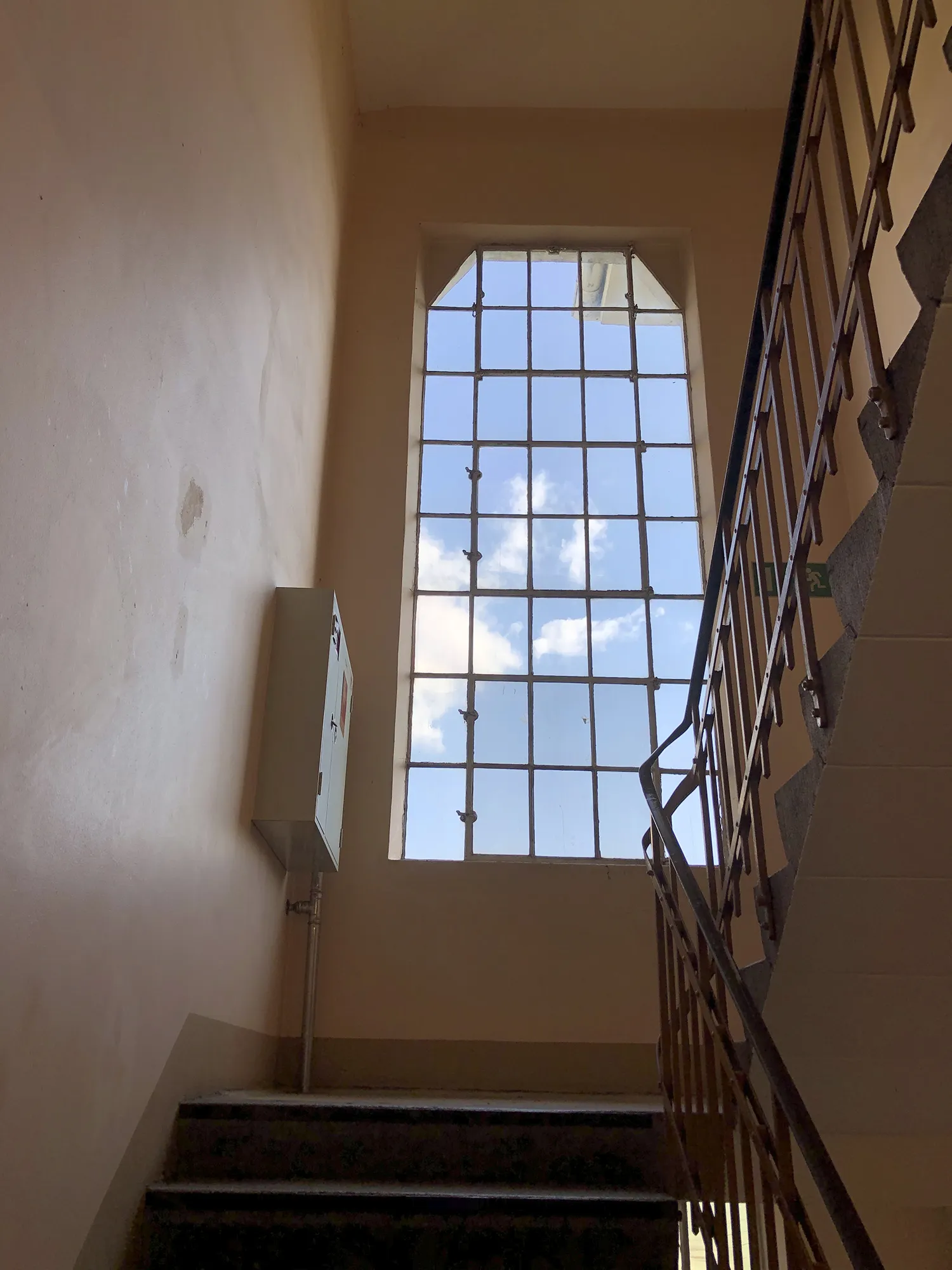Treppenhaus mit Metallgeländer und großem Sprossenfenster, durch das blauer Himmel mit weißen Wolken sichtbar ist, an der Wand ein kleiner Verteilerkasten.