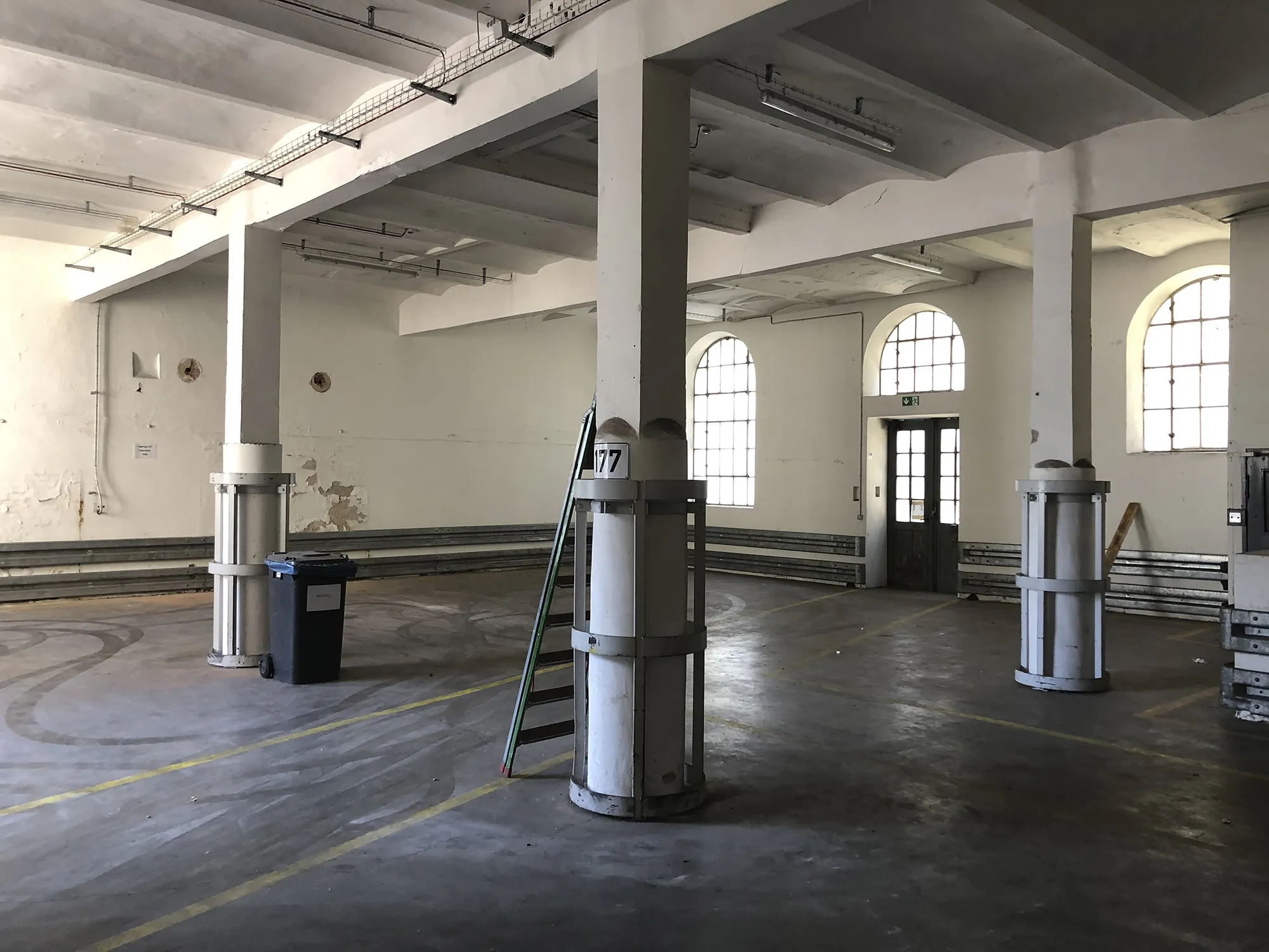 Leere Industriehalle mit weißen Stützsäulen, hohen Bogenfenstern, auf dem Boden gelbe Markierungen, im Vordergrund eine Leiter und ein Abfallbehälter.