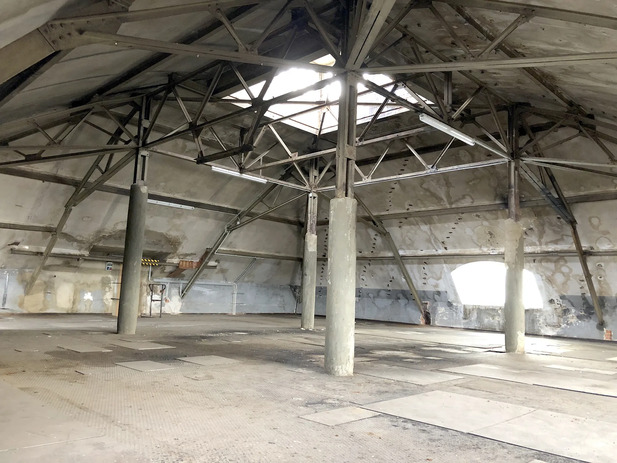 Innenansicht eines großen, leeren Dachbodens mit Betonstützen, Metallträgern und abgenutzten Wänden, ganz obn ein Deckenfenster mit Lichteinfall.