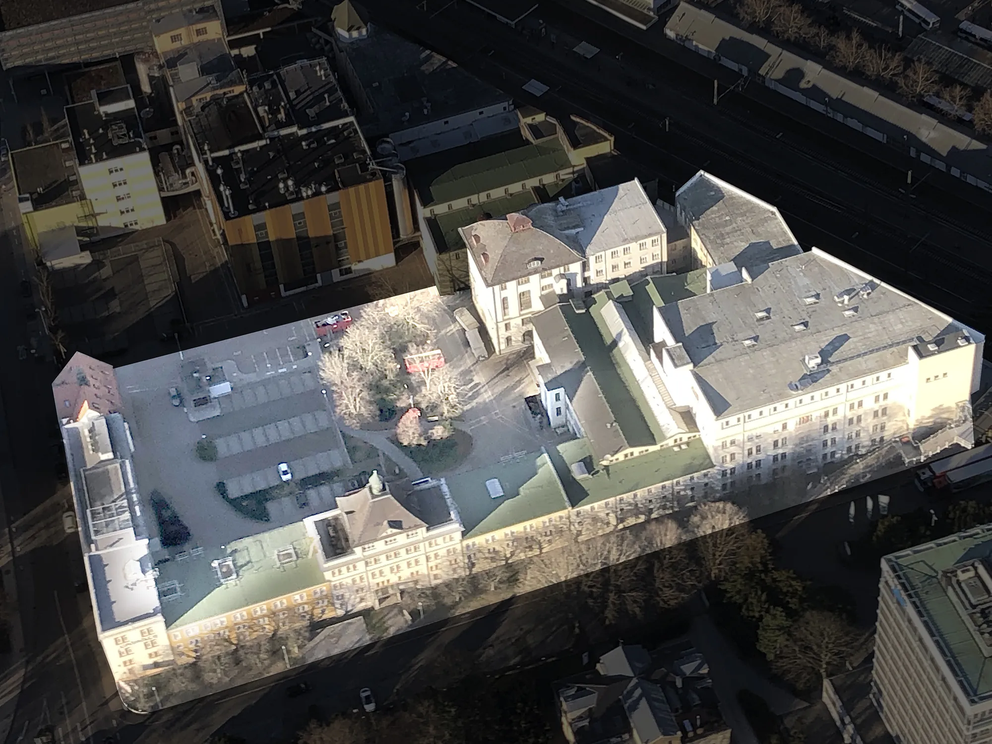 Luftaufnahme eines hervorgehobenen großen Gebäudekomplexes (Caro Carré) mit mehreren Flügeln und Innenhof, umgeben von Straßen, Parkplätzen und Bäumen, im Hintergrund Bahngleise und angrenzende Stadtbebauung.