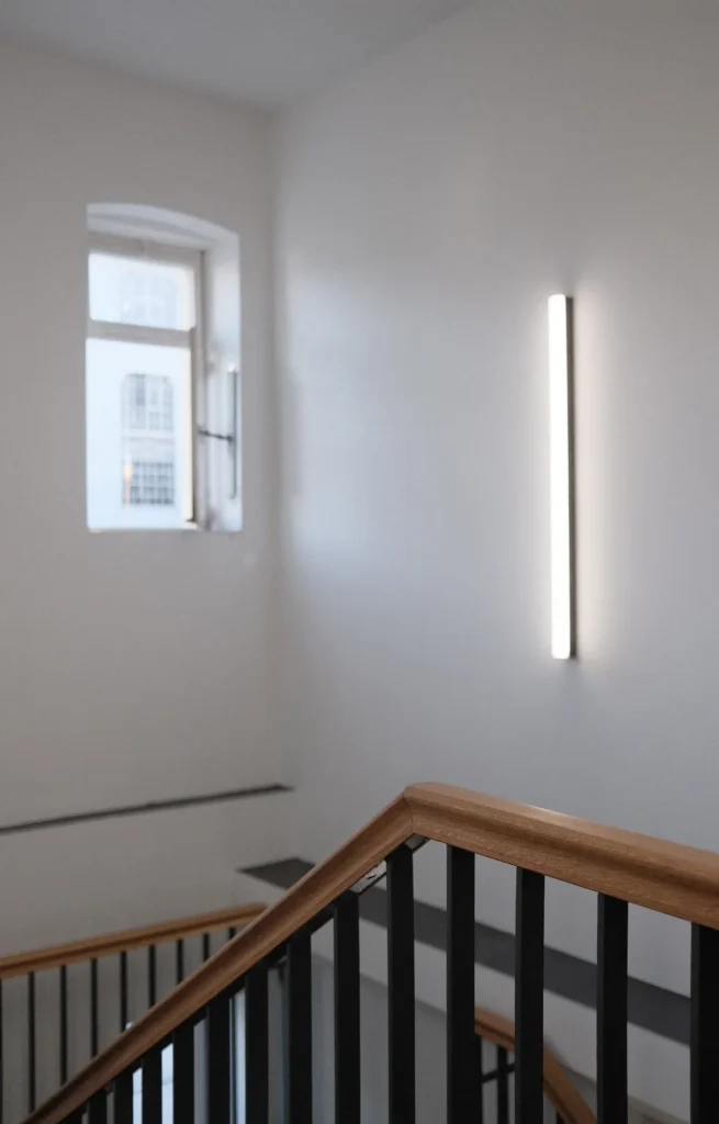 Treppenhaus mit schwarzem Geländer und hölzernem Handlauf, im Hintergrund ein schmales, hochkant angebrachtes Wandleuchtobjekt und ein geöffnetes Fenster, durch das ein Teil eines gegenüberliegenden Gebäudes sichtbar ist.
