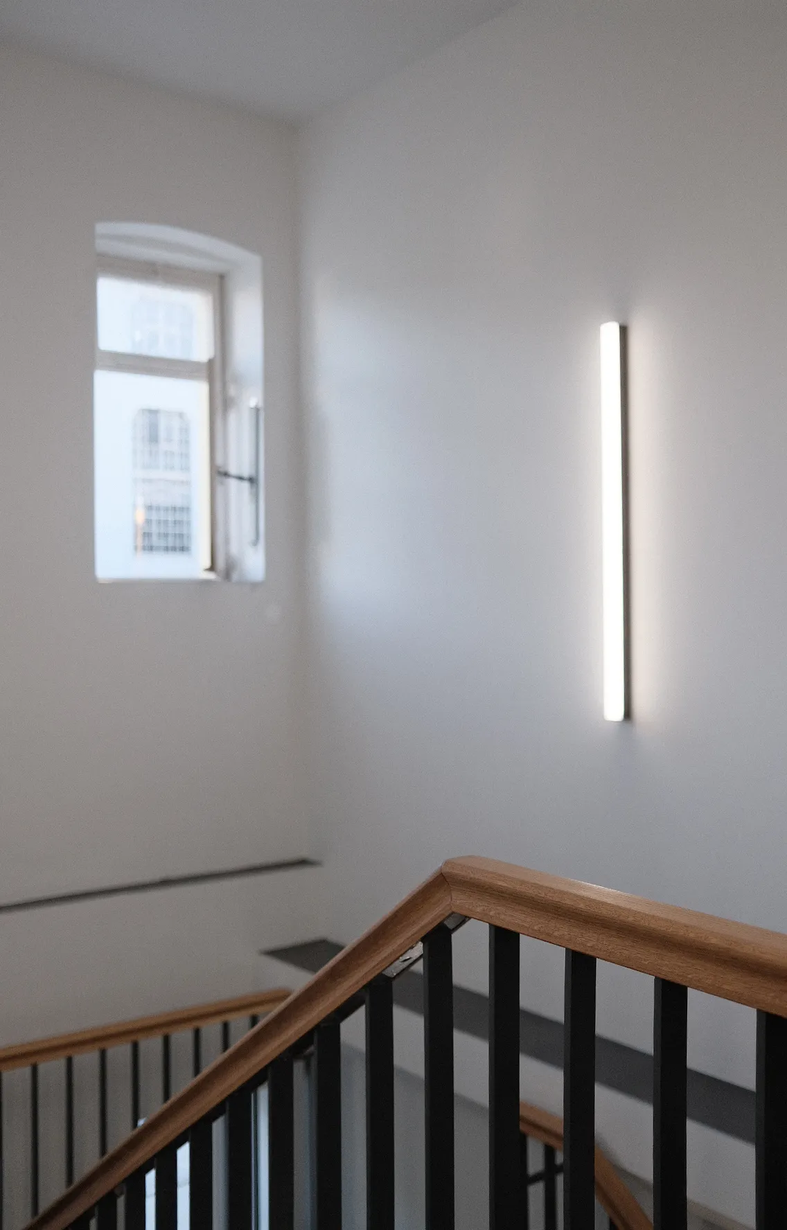 Treppenhaus mit schwarzem Geländer und hölzernem Handlauf, im Hintergrund ein schmales, hochkant angebrachtes Wandleuchtobjekt und ein geöffnetes Fenster, durch das ein Teil eines gegenüberliegenden Gebäudes sichtbar ist.
