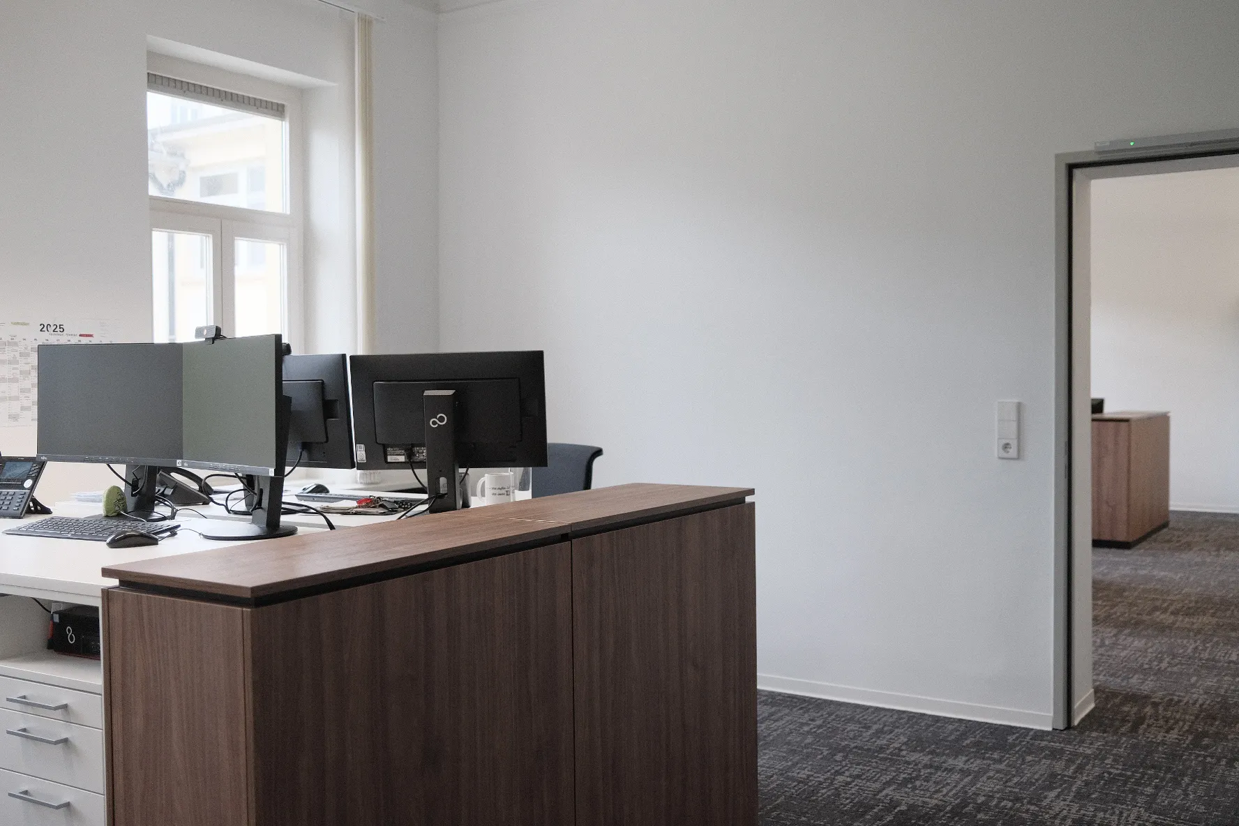 Modernes Büro mit zwei Arbeitsplätzen, jeweils ausgestattet mit mehreren Monitoren und Telefonen, auf weißen Schreibtischen. Im Vordergrund ein hoher Schrank mit Holzoptik, im Hintergrund ein Durchgang zu einem weiteren Raum mit ähnlicher Büroeinrichtung.