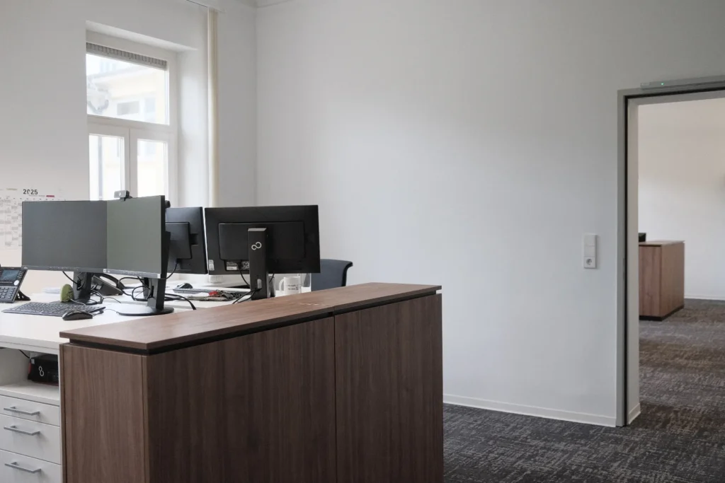 Modernes Büro mit zwei Arbeitsplätzen, jeweils ausgestattet mit mehreren Monitoren und Telefonen, auf weißen Schreibtischen. Im Vordergrund ein hoher Schrank mit Holzoptik, im Hintergrund ein Durchgang zu einem weiteren Raum mit ähnlicher Büroeinrichtung.