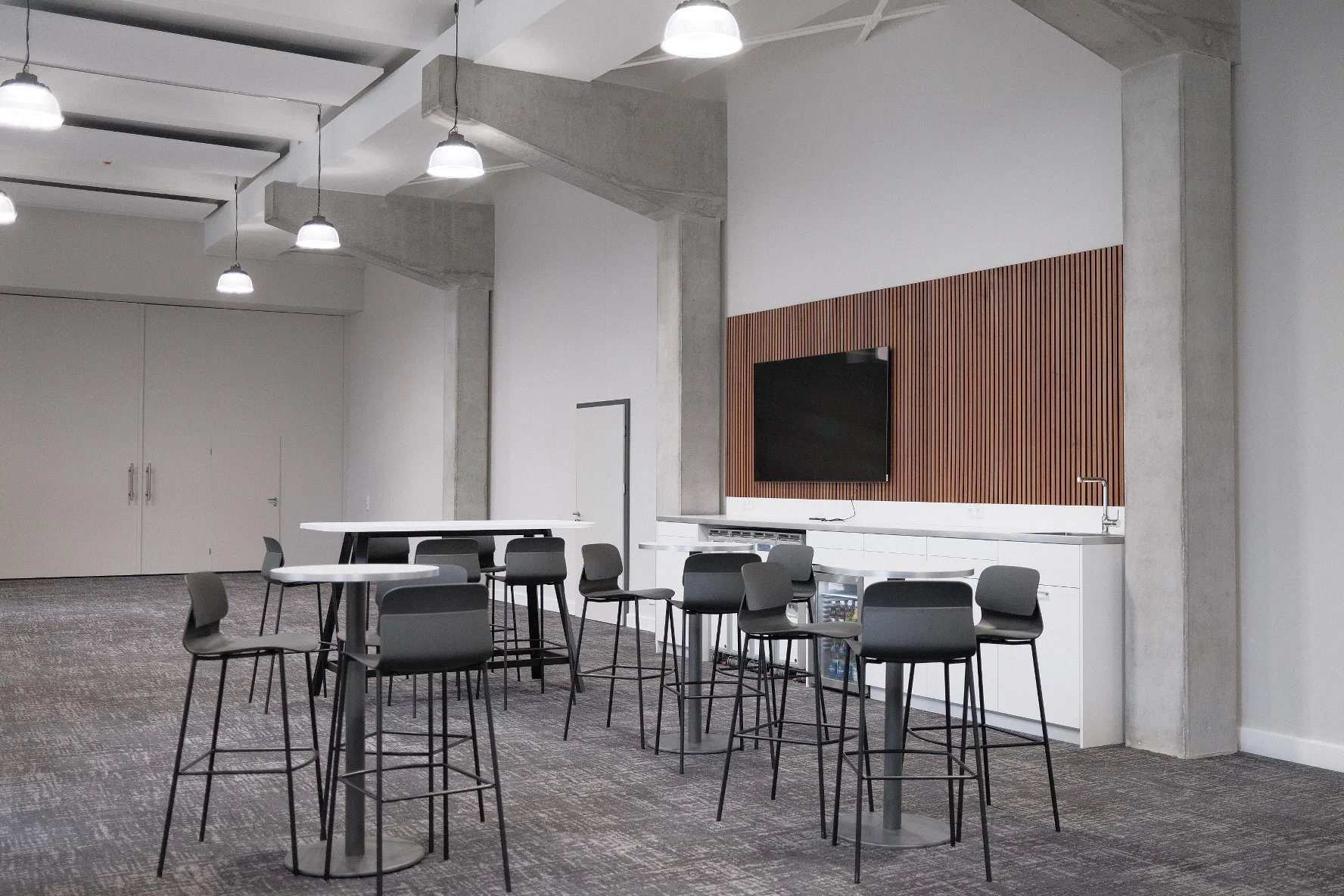 Moderner Aufenthaltsbereich mit hohen Decken, grauem Teppichboden und Betonstützen. Mehrere hohe Tische mit Barhockern stehen locker verteilt, an der Rückwand eine weiße Küchenzeile mit eingebautem Kühlschrank, Spülbecken und einer hölzernen Wandverkleidung mit Flachbildfernseher. Hängende Industrieleuchten beleuchten den Raum.