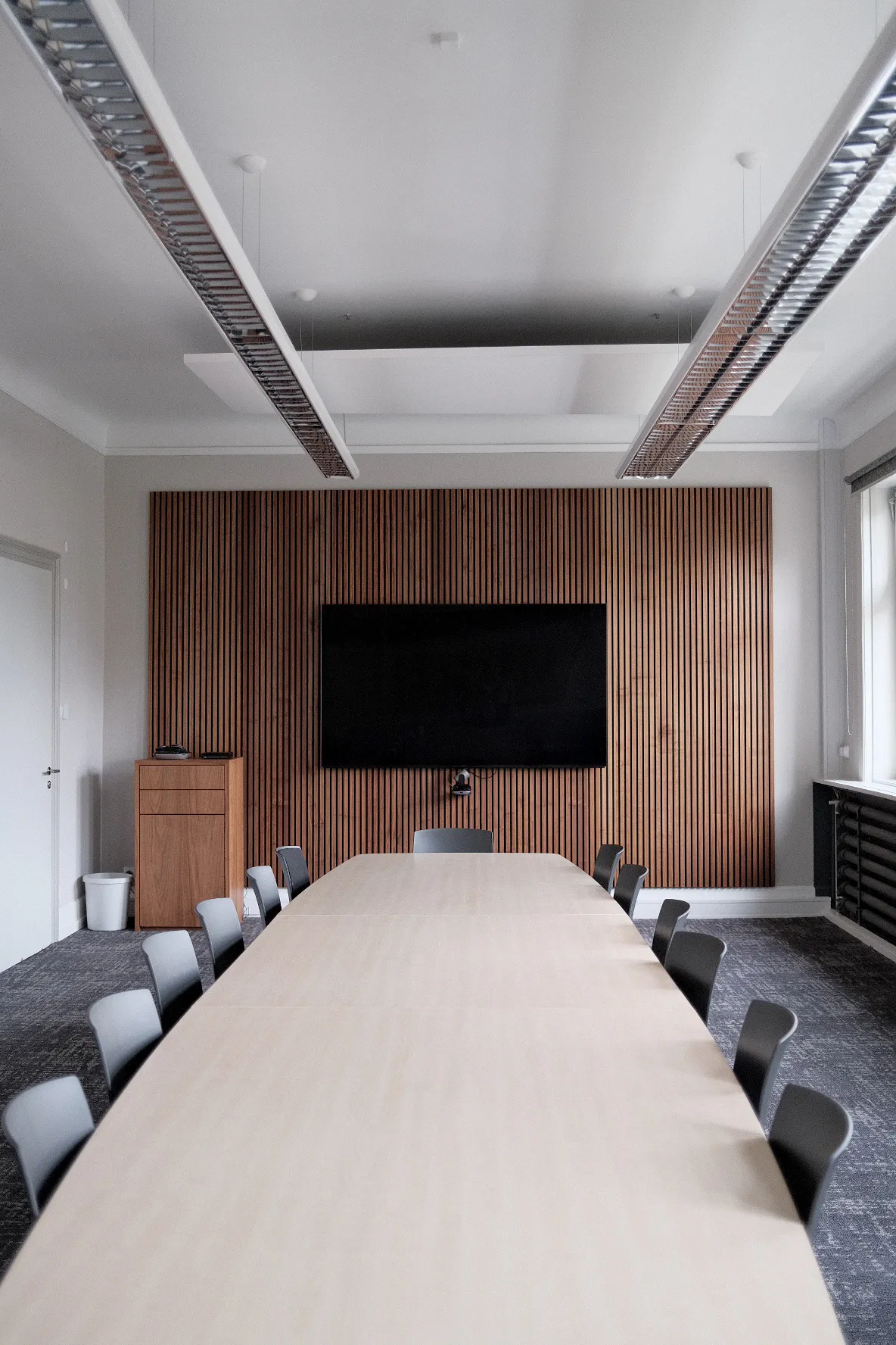 Moderner Konferenzraum mit großem rechteckigen Holztisch, umgeben von grauen Stühlen. An der Stirnseite eine Wand mit vertikaler Holzverkleidung und großem Flachbildschirm, darunter eine Kamera. Links steht ein kleines Rednerpult, rechts fällt Tageslicht durch ein Fenster. An der Decke sind lange, schmale Hängelampen installiert.