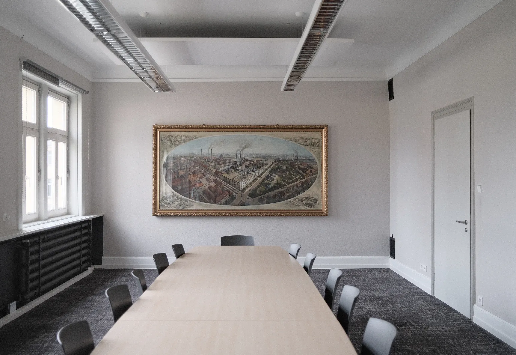 Konferenzraum mit großem rechteckigen Holztisch, umgeben von grauen Stühlen. An der Stirnseite hängt ein großes gerahmtes Panoramagemälde einer historischen Industrieanlage an einer hellgrauen Wand. Links fällt Tageslicht durch zwei große Fenster, rechts befindet sich eine geschlossene Tür. An der Decke sind lange Hängelampen installiert.
