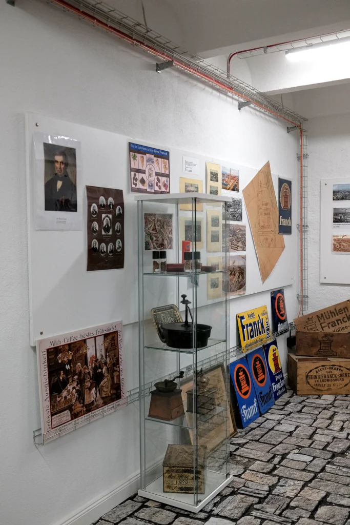 In einem Ausstellungsraum steht im Vordergrund eine Vitrine mit alten Gerätschaften wie zum beispiel einer Kaffeemühle. Ringsherum hängen Bilder und Aufzeichnungen an einem Whiteboard.