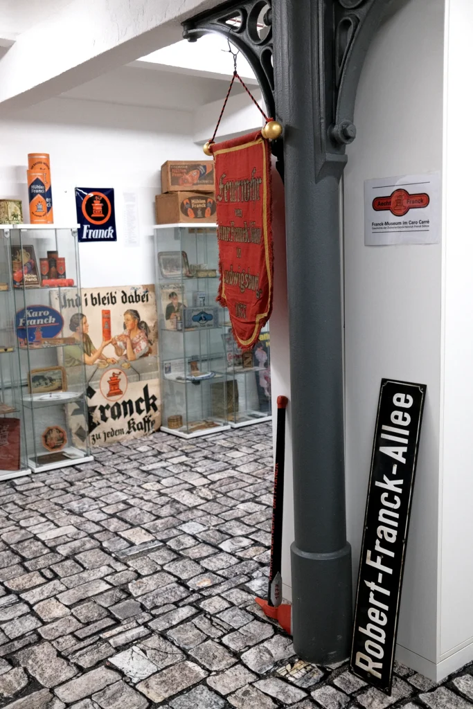 Fotografie in einen Austellungsraum. Im Vordrgrund stejt ein altes Straßenschild an einen Schrank gelehnt mit der Aufschrift "Robert-Franck-Allee". Daneben hängt ein Wimpel der Feuerwehr von Heinr. Franck von einer Säule . Im Hintergrund stehen Vitrinen und Schilder an der Wand.