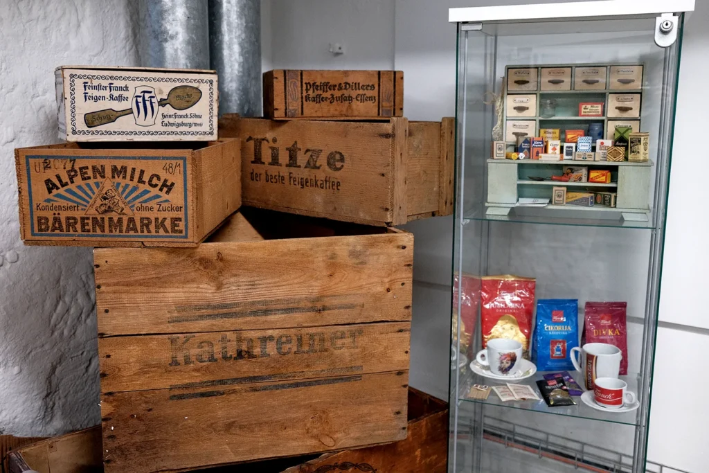 In einer Raumecke sind mehrere Holzkisten aufgestellt. Eine trägt die Aufschrift "Titze" eine andere "Kathreiner". neben den Kisten ist die obere Hälfte einer Vitrine zu sehen, die einen miniatur Modellladen sowie Produkte der Firma Frank enthält. Darunter Kaffeepackungen und Tassen.