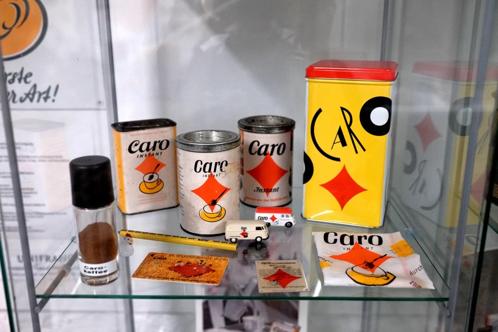 Nahaufnahme einers Vitrinenfaches in dem verschiedene (Werbe-) Produkte der Marke Caro ausgestellt sind. Darunter ein paar Kaffeedosen, ein Bleistift, zwei Modellautos und ein Tuch. Alles ist in den Farben weiß, gelb, rot-orange mit schwarzer Schrift gehalten