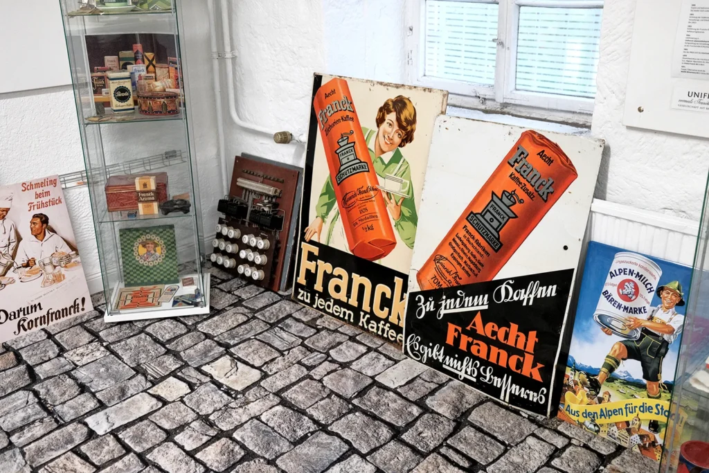 Plakate und Schilder der Firma Aecht Franck sowie Bärenmarke sind an eine Wand gelehnt. Auf den präsentesten sind große orangene Kaffee-Rollen mit der Aufschrift "Aecht Franck Kaffee-Zusatz" und einer Kaffeemühlengrafik zu sehen.
