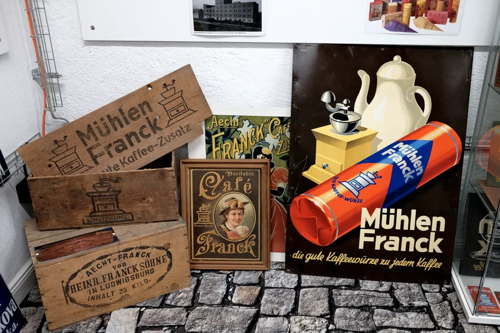 Plakate und Schilder der Firma Mühlen Franck sind an eine weiße Wand gelehnt. Eines davon zeigt zum Beispiel im Hintergrund eine weiße Kaffeekanne auf schwarzer Fläche, davor eine haushaltsübliche Kaffeemühle und davor eine Rolle Kaffee. Neben den Schildern stehen Holzkisten und -Schilder mit der Aufschrift "Mühlen Franck".