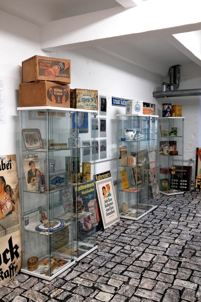 Ein heller Raum mit mehreren Vitrinen die Produkten einer ehemaligen Kaffe-Rösterei austellen. Zwischen den Vitrinen stehen und hängen verschiedene Werbeplakate und Schilder an der Wand.