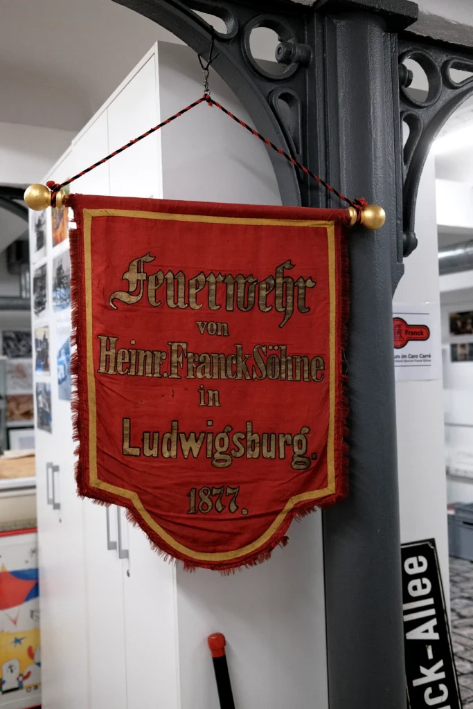 Ein roter Wimpel mit goldener Verzierung und der Aufschrift "Feuerwehr von Heinr. Franck Söhne in Ludwigsburg. 1877." hängt von einem Pfeiler.