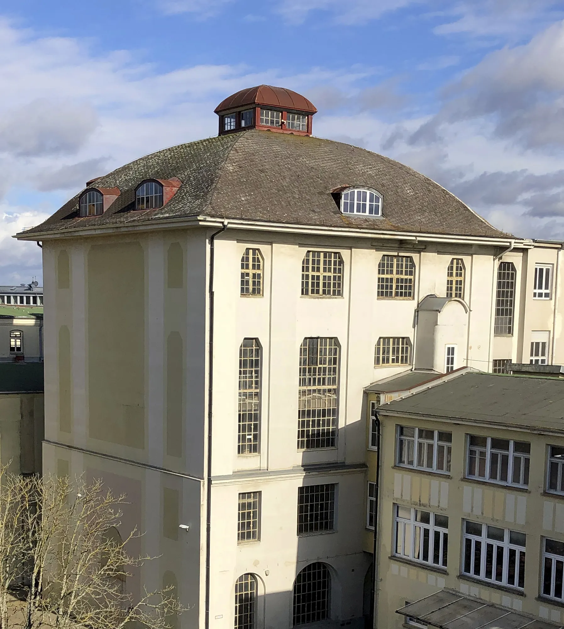 Historisches mehrstöckiges Gebäude mit heller Fassade, großen Bogen- und Sprossenfenstern sowie einem gewölbten Dach mit rotem Dachaufbau, vor blauem Himmel mit Wolken.