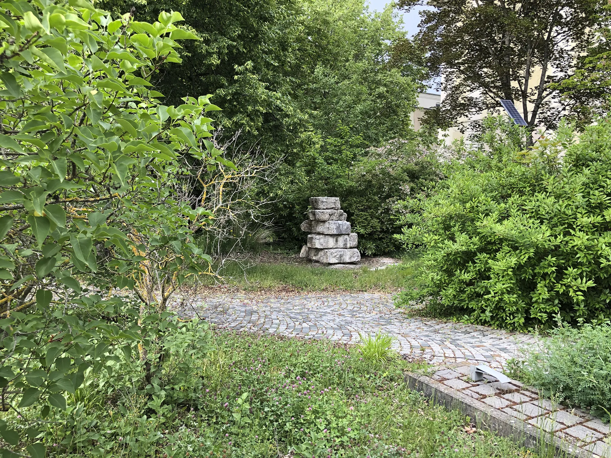 Gartenbereich mit gepflastertem Weg, umgeben von dichtem Grün und Büschen, im Hintergrund ein kleiner Stapel aus groben Steinblöcken.