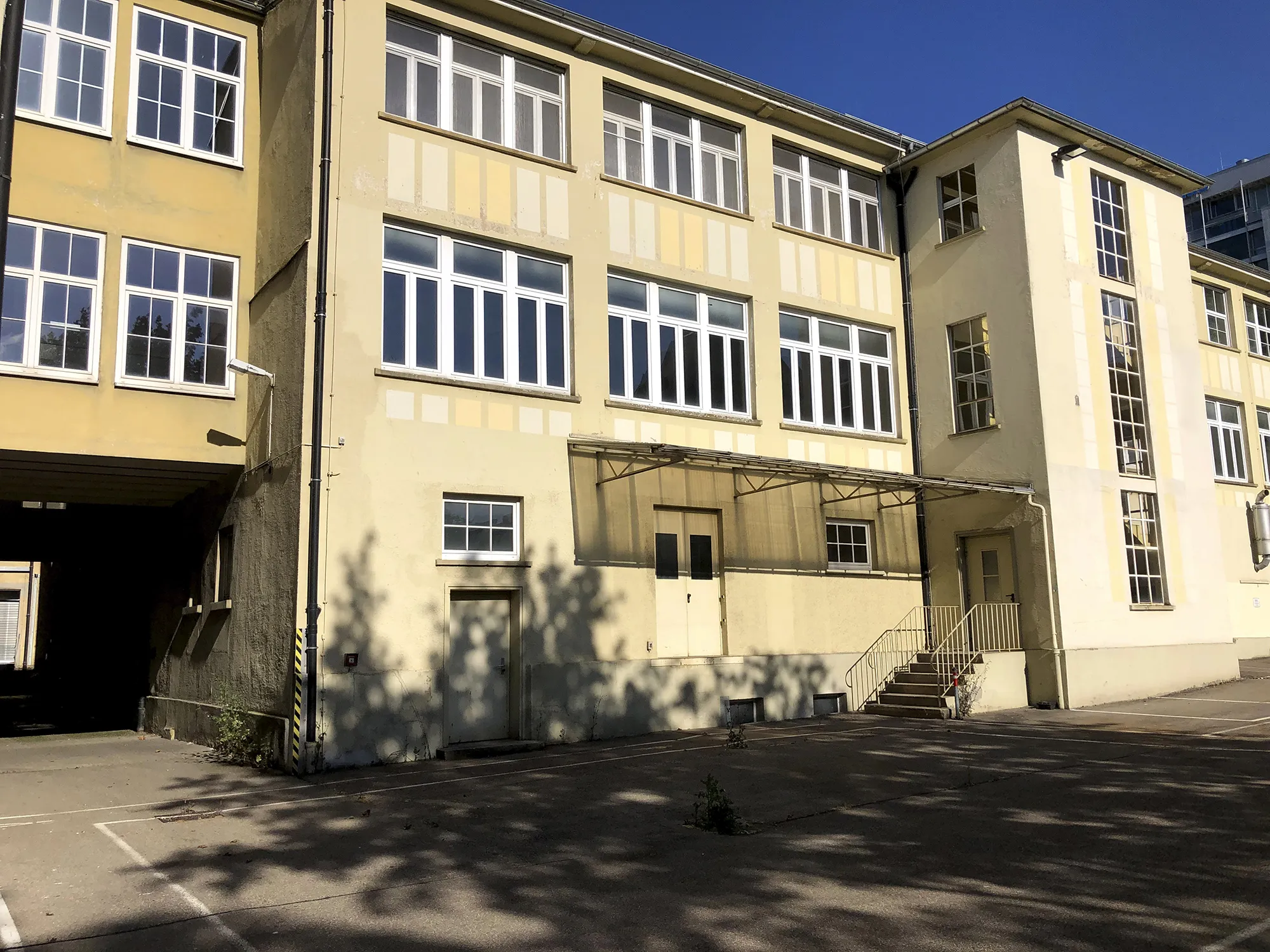Fassade eines hellgelben mehrstöckigen Gebäudes mit großen weißen Fenstern, Treppe zum Eingang auf der rechten Seite und überdachtem Bereich über einer Doppeltür, links ein Durchgang zu einem Innenhof.