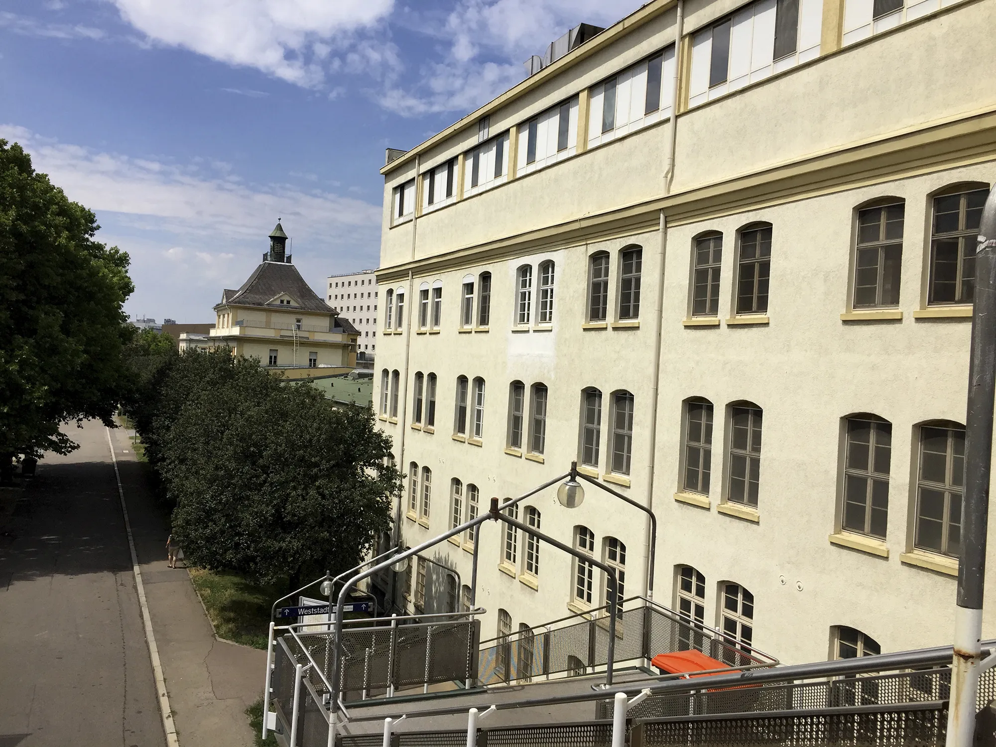 Blick auf eine lange, beige Fassade eines mehrstöckigen Gebäudes mit vielen Fenstern, im Hintergrund ein weiteres Gebäude mit Turmspitze und grünem Dach, links gesäumt von einer Baumreihe an einer Straße.