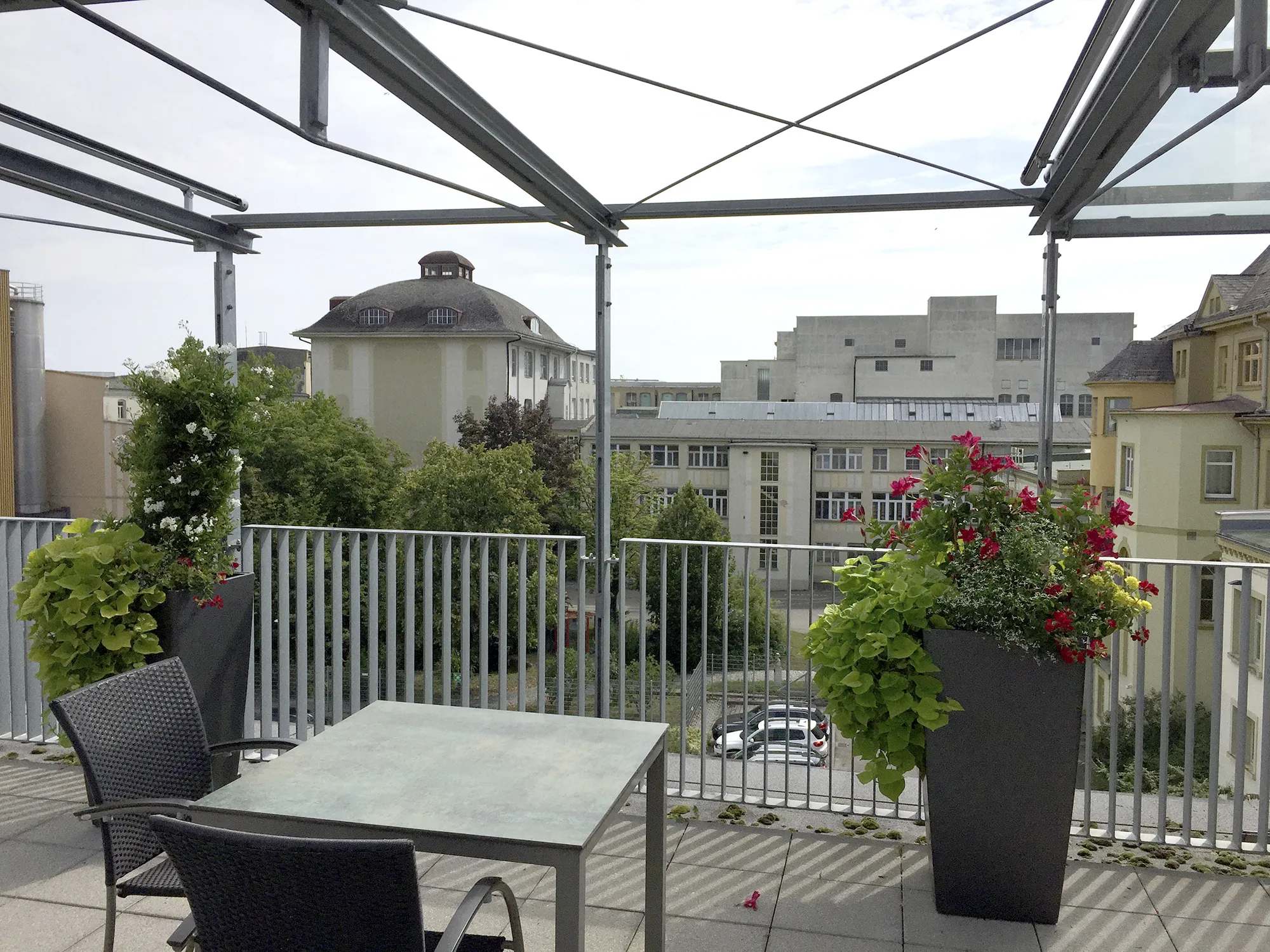 Dachterrasse mit Tisch und Stühlen, flankiert von großen Blumentöpfen mit roten und weißen Blüten, im Hintergrund Blick auf Bäume und umliegende Gebäude.