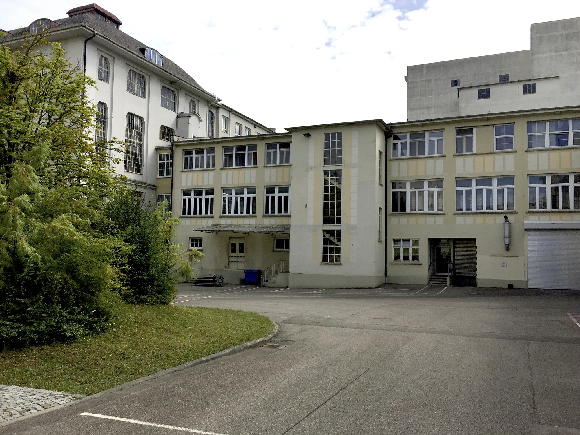 Ansicht eines mehrstöckigen Industrie- und Bürogebäudes mit hellgelber und weißer Fassade, großen Sprossenfenstern und einem begrünten Vorbereich, im Vordergrund ein leerer asphaltierter Parkplatz.